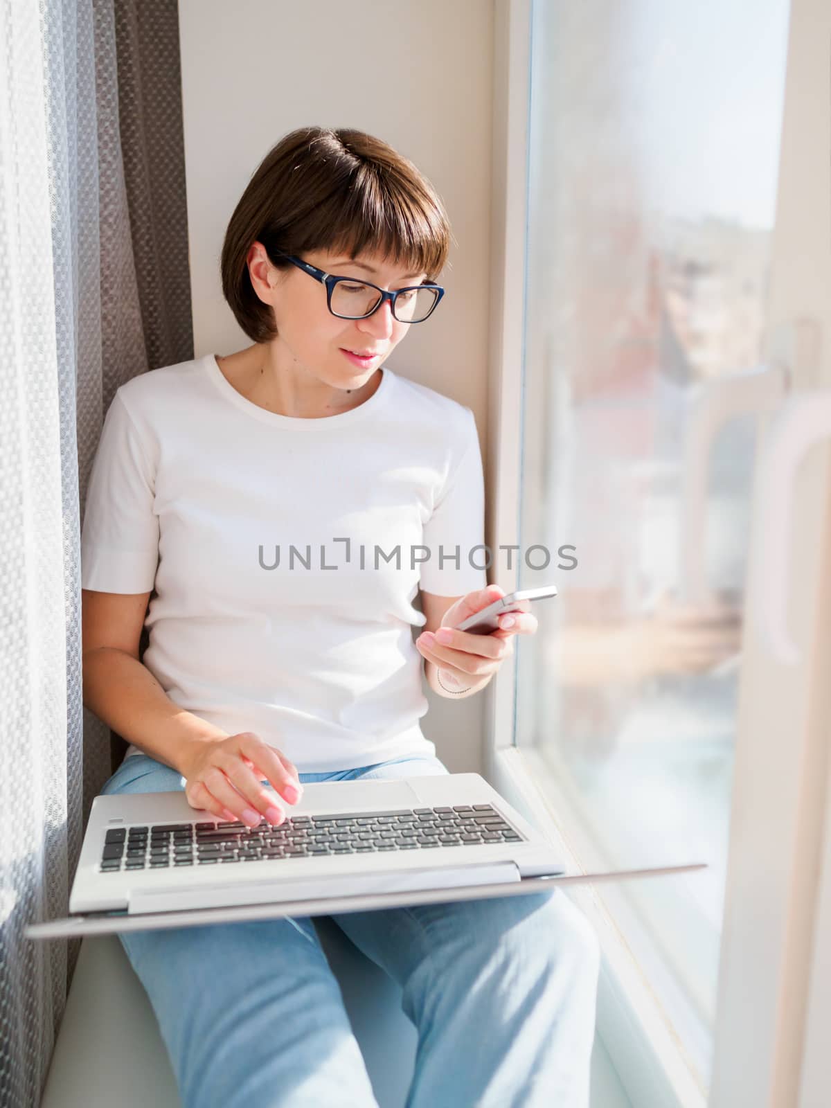 Woman works remotely from home. She sits on window sill with lap by aksenovko