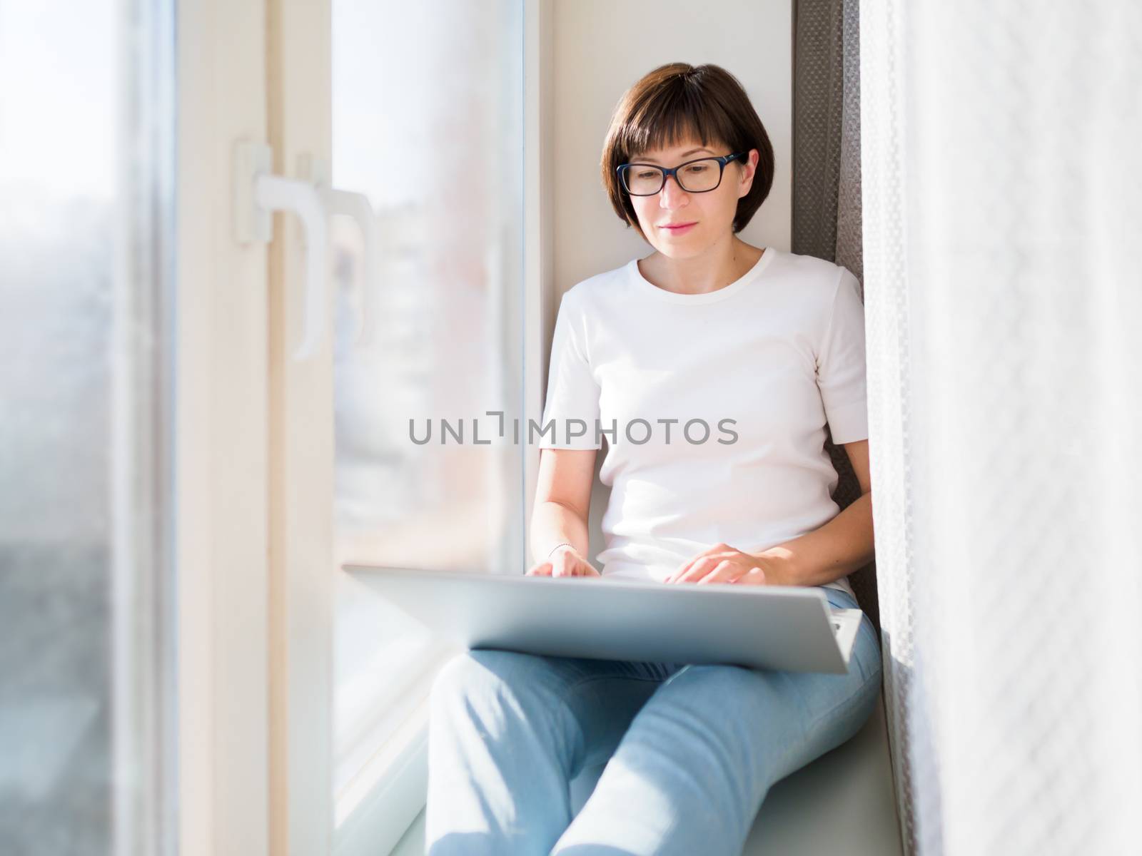 Woman works remotely from home. She sits on window sill with laptop on knees. Lockdown quarantine because of coronavirus COVID19. Self isolation at home.