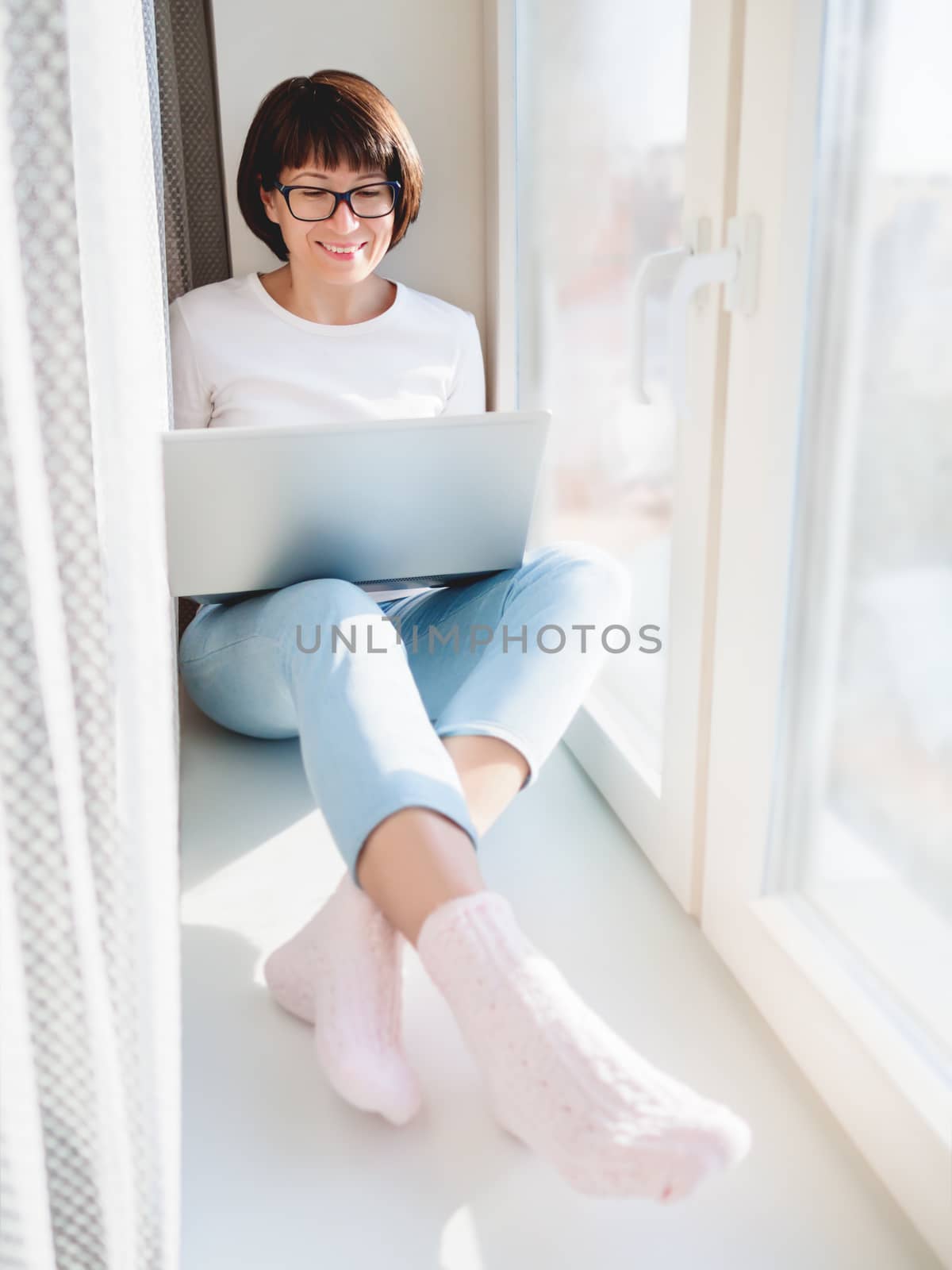 Smiling woman works remotely from home. She sits on window sill with laptop on knees. Lockdown quarantine because of coronavirus COVID19. Self isolation at home.