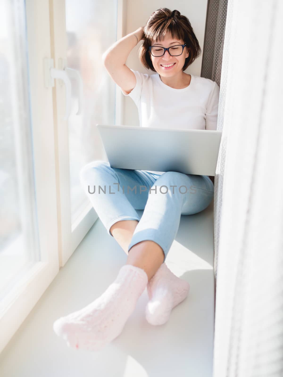 Smiling woman works remotely from home. She sits on window sill by aksenovko