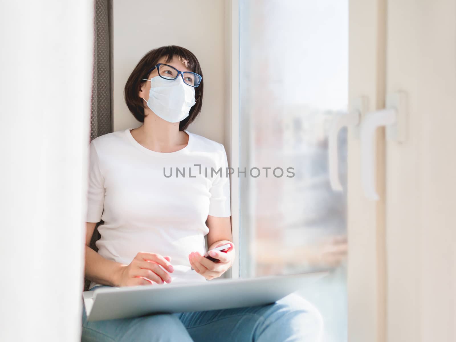 Woman in medical protective mask works remotely from home. She sits on window sill with smartphone and laptop on knees. Lockdown quarantine because of coronavirus COVID19.