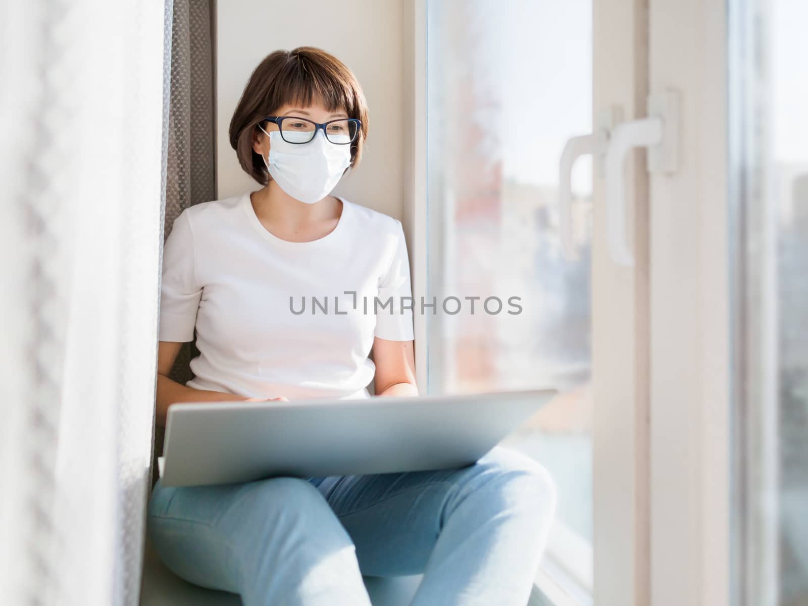 Pretty woman in medical mask works remotely from home. She sits on window sill with laptop on knees. Lockdown quarantine because of coronavirus COVID19. Self isolation at home.