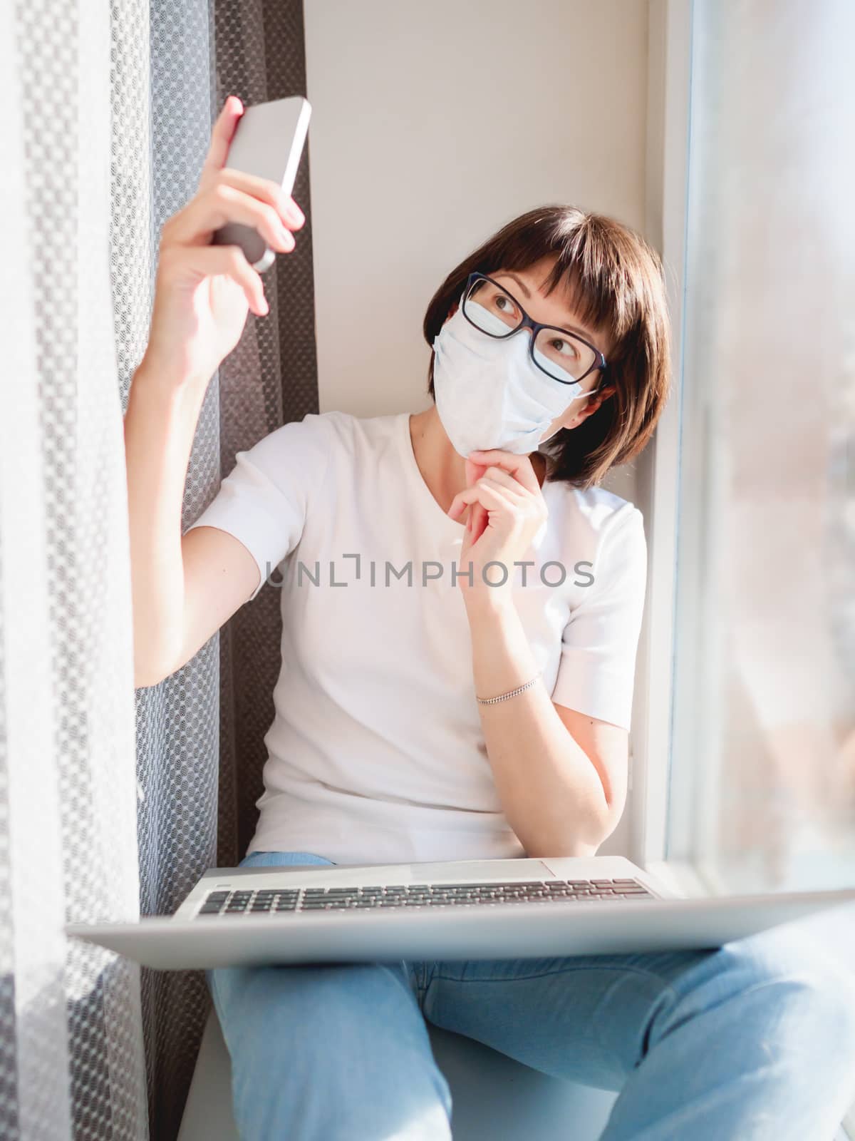 Pretty woman in medical mask works remotely from home. She is making selfie on window sill with laptop on knees. Lockdown quarantine because of coronavirus COVID19. Self isolation at home.