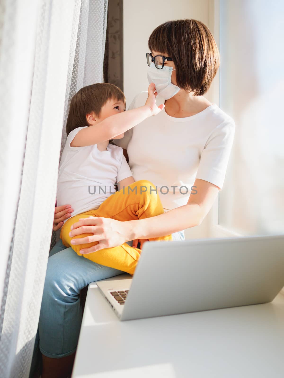 Woman in protective medical mask works remotely from home. She sits on window sill with laptop and cute toddler boy on her knees. Lockdown quarantine because of coronavirus COVID19. Self isolation.