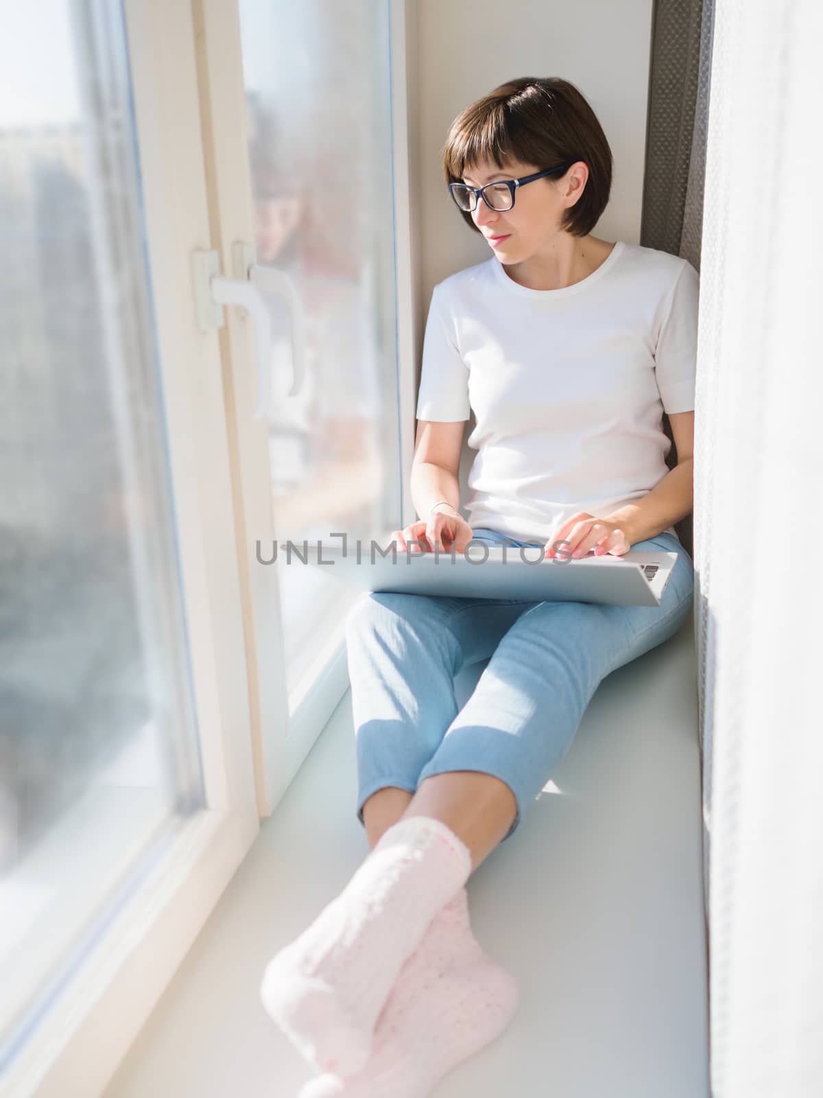 Woman works remotely from home. She sits on window sill with laptop on knees. Lockdown quarantine because of coronavirus COVID19. Self isolation at home.