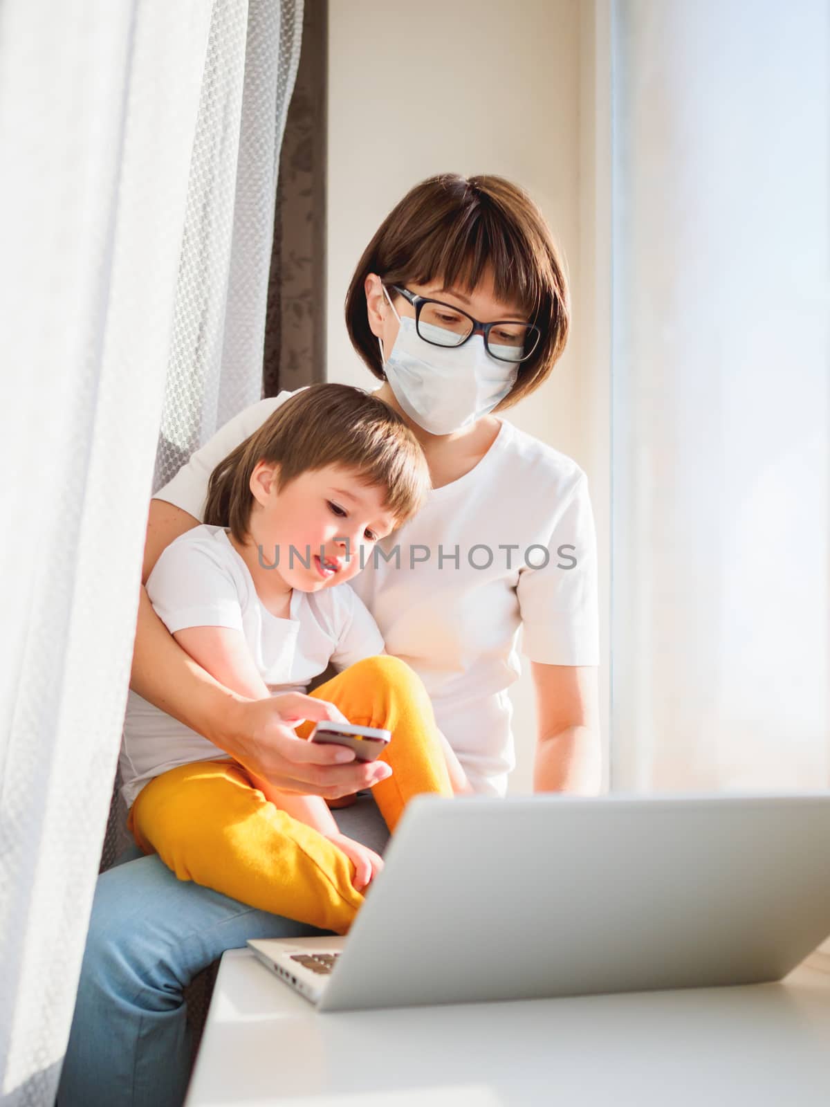 Woman in protective medical mask works remotely from home. She sits on window sill with laptop and cute toddler boy on her knees. Lockdown quarantine because of coronavirus COVID19. Self isolation.