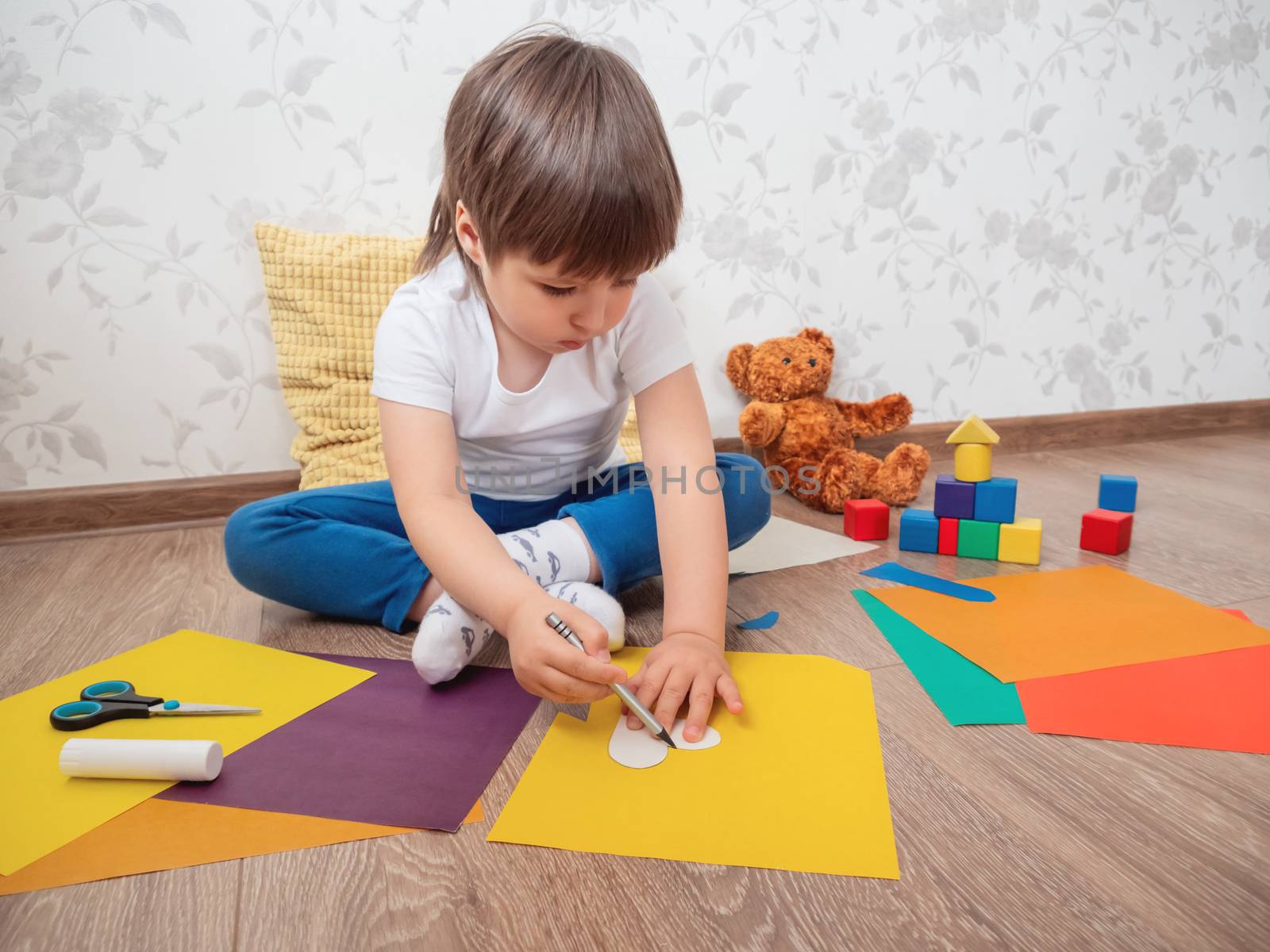 Toddler boy learns to cut colored paper with scissors. Kid sits on floor in kids room with toy blocks and teddy bear. Educational classes for children. Developing feeling sensations and fine motor skills at home.