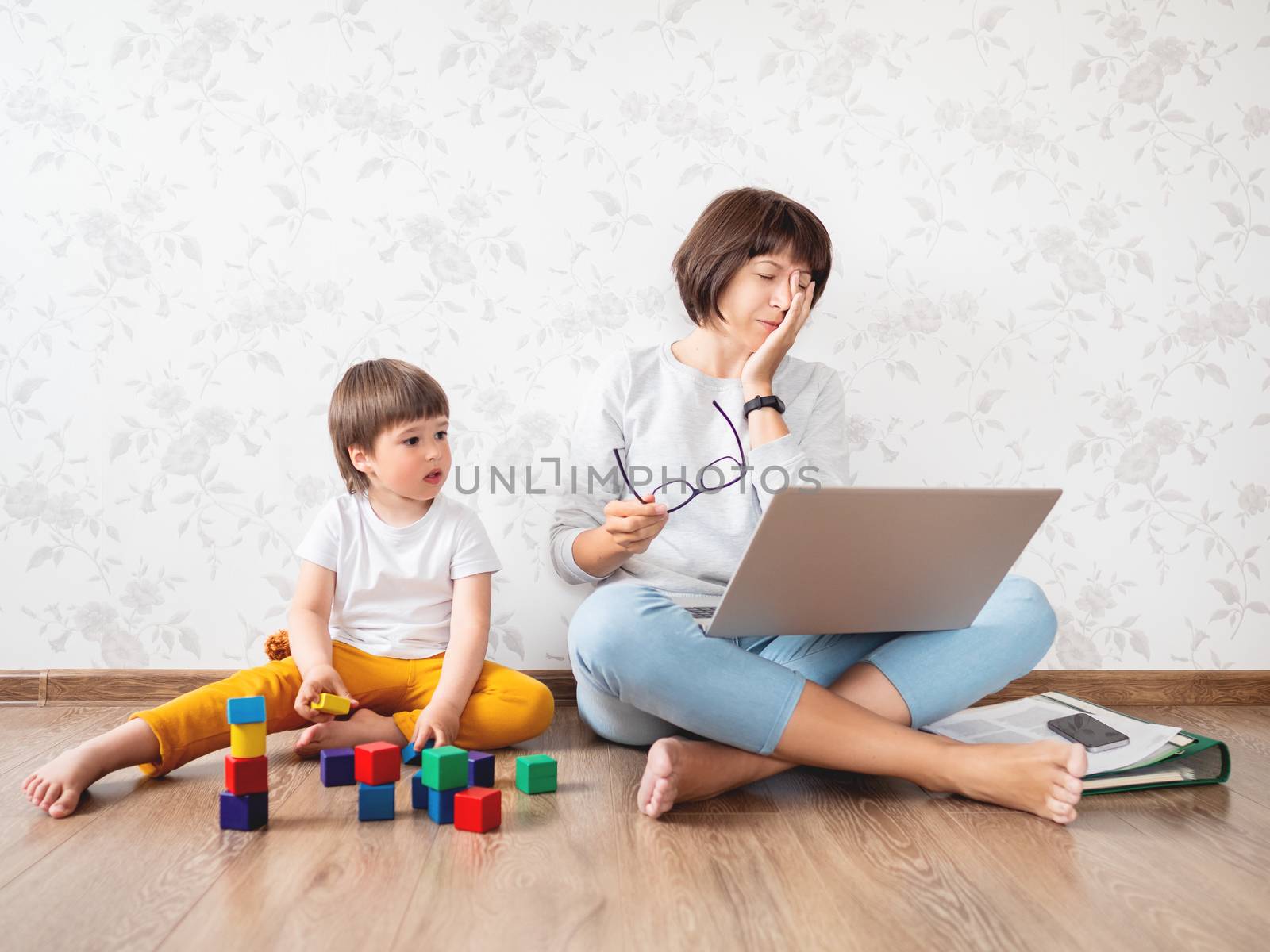 Mom and son argue at home quarantine because of coronavirus COVID19. Tired mother works remotely with laptop, son plays with toy blocks. Self isolation at home.