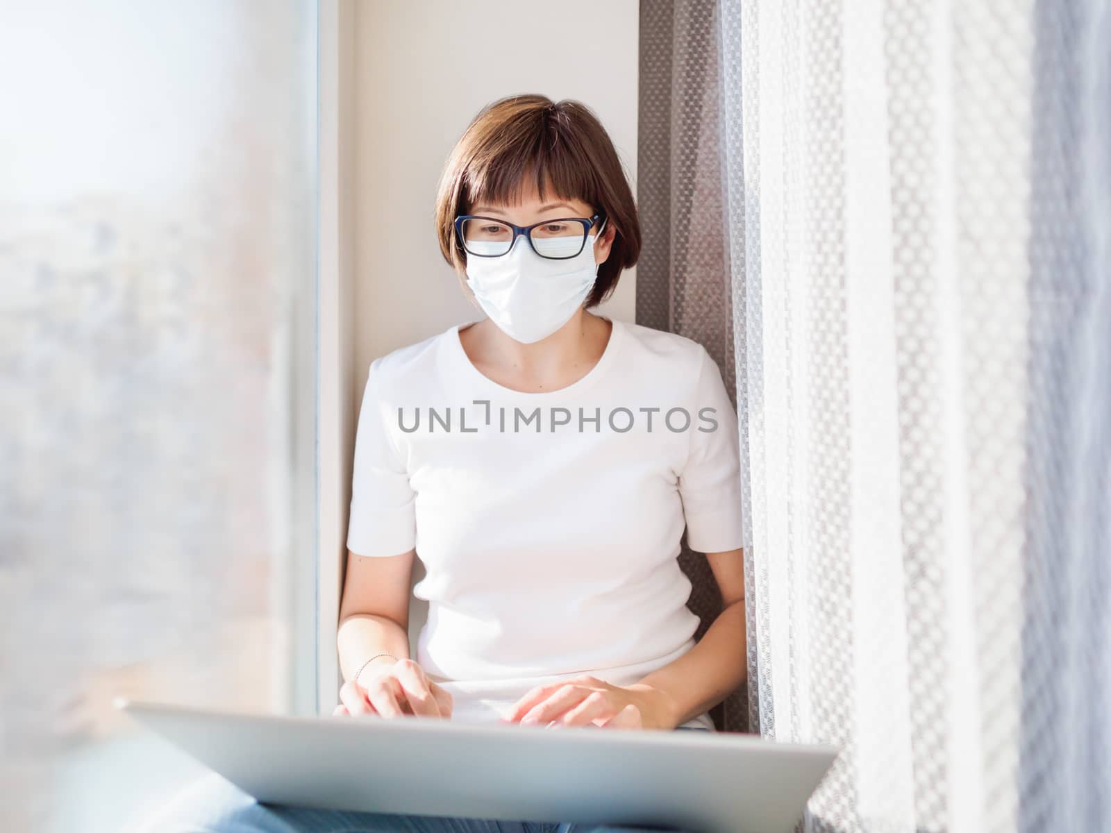 Pretty woman in medical mask works remotely from home. She sits on window sill with laptop on knees. Lockdown quarantine because of coronavirus COVID19. Self isolation at home.