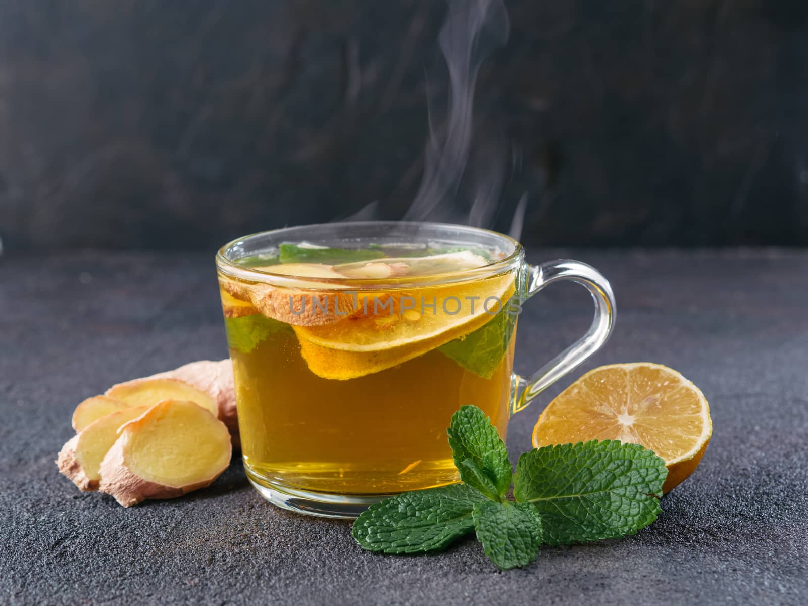 Herbal tea with ginger, mint and lemon. Hot tea glass cup with steam and ingredients on black cement background. Copy space