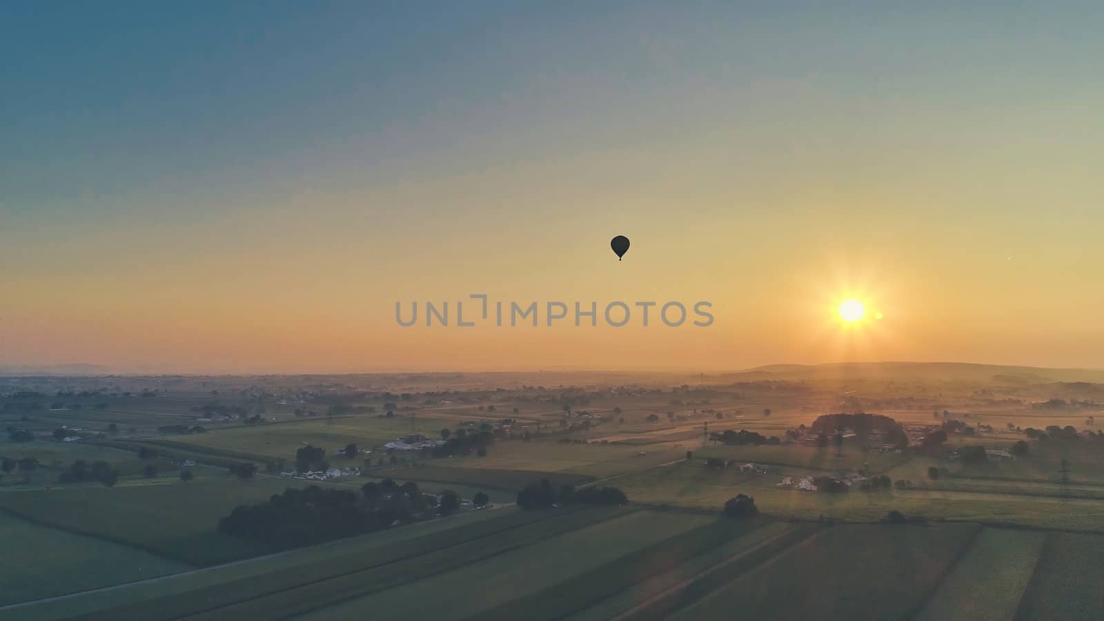 Aerial view of a summer sunset over farm lands with blue skies and red sun and a hot air balloon floating by