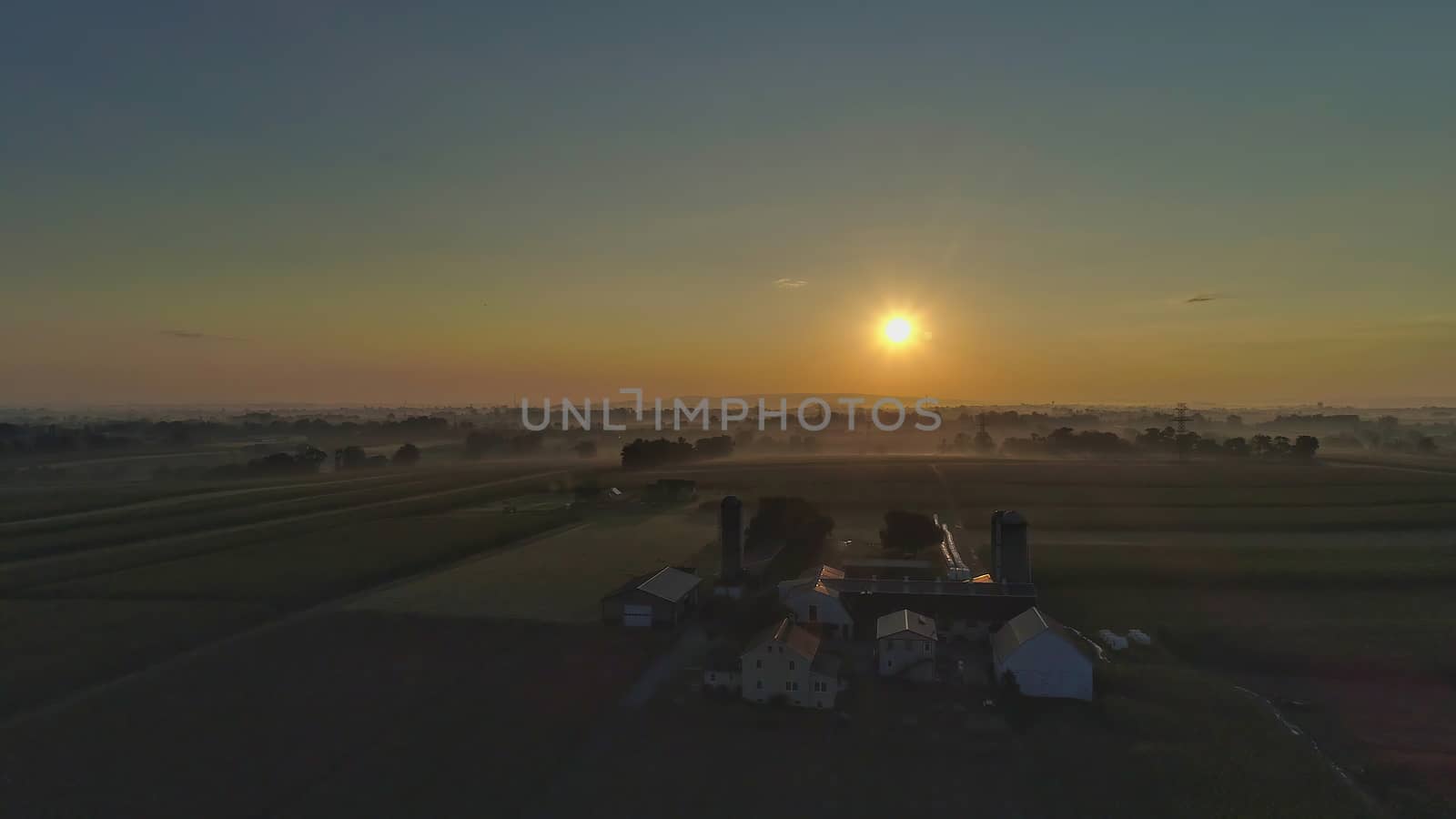 Sunrise over barns, silos and green farmlands with blue, red and orange skies and a few clouds with a mist on the ground