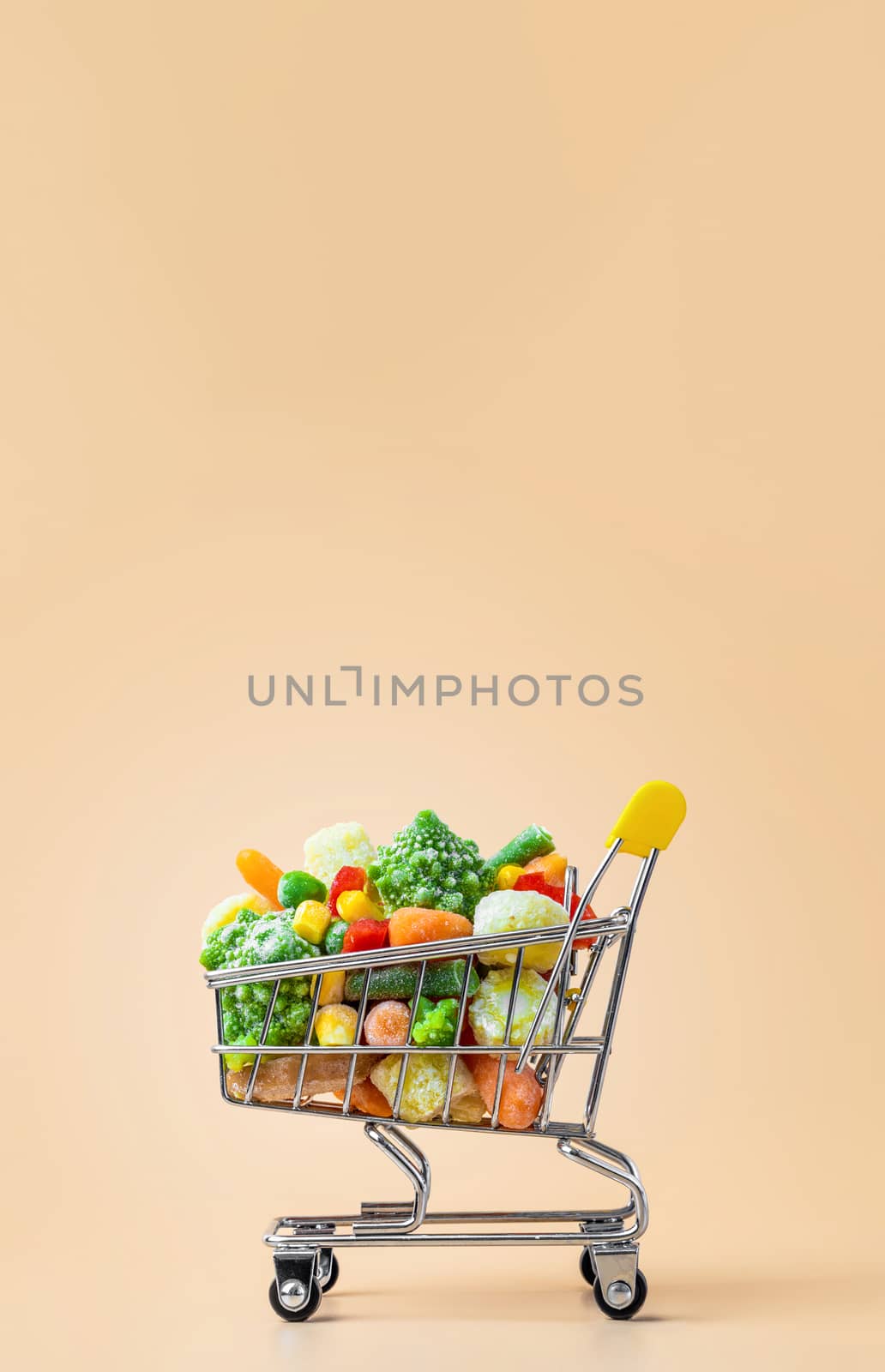 Frozen vegetables assorted in toy shopping cart on cream background. Full of assorted frozen vegetables food shop trolley at beige or yellow backdrop. Minimalistic concept. Vertical. Copy space