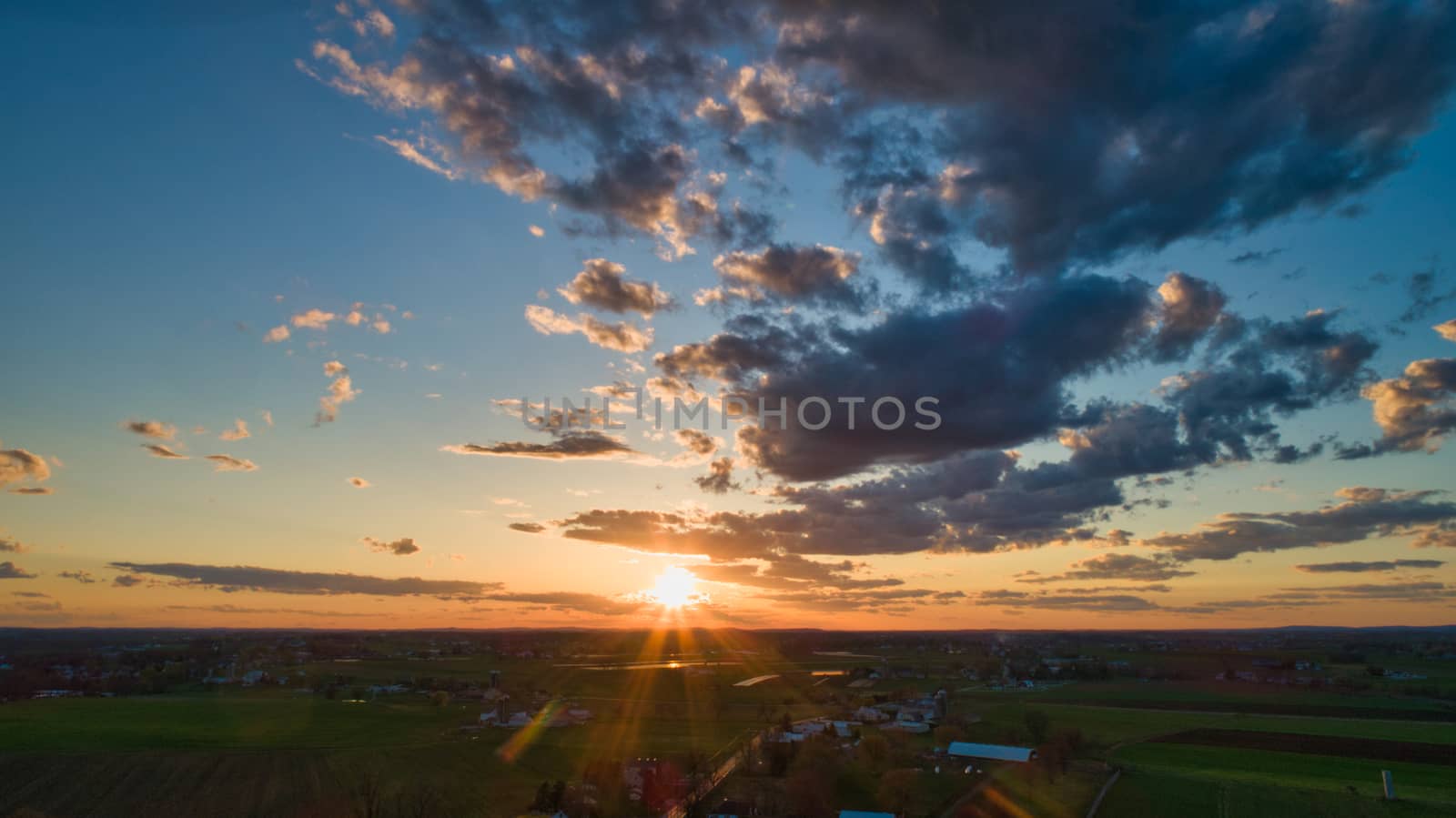Aerial view of a Sunset over barns, silos and farmlands during the golden hour with blue sky, clouds and red sun
