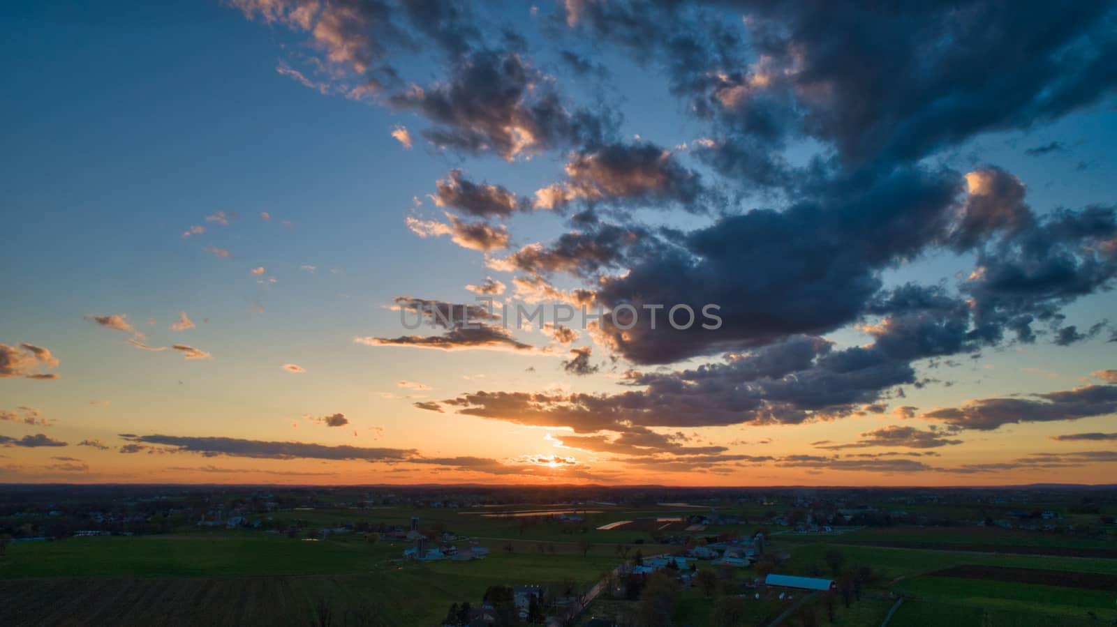 Aerial view of a Sunset over barns, silos and farmlands during the golden hour with blue sky, clouds and red sun