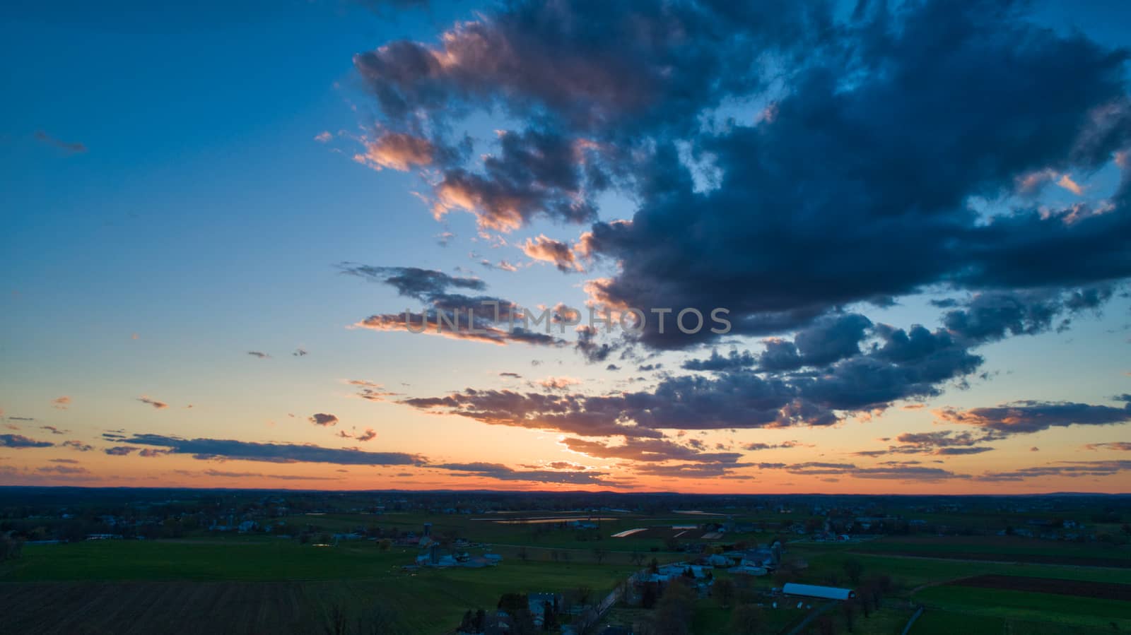 Aerial view of a Sunset over barns, silos and farmlands during the golden hour with blue sky, clouds and red sun