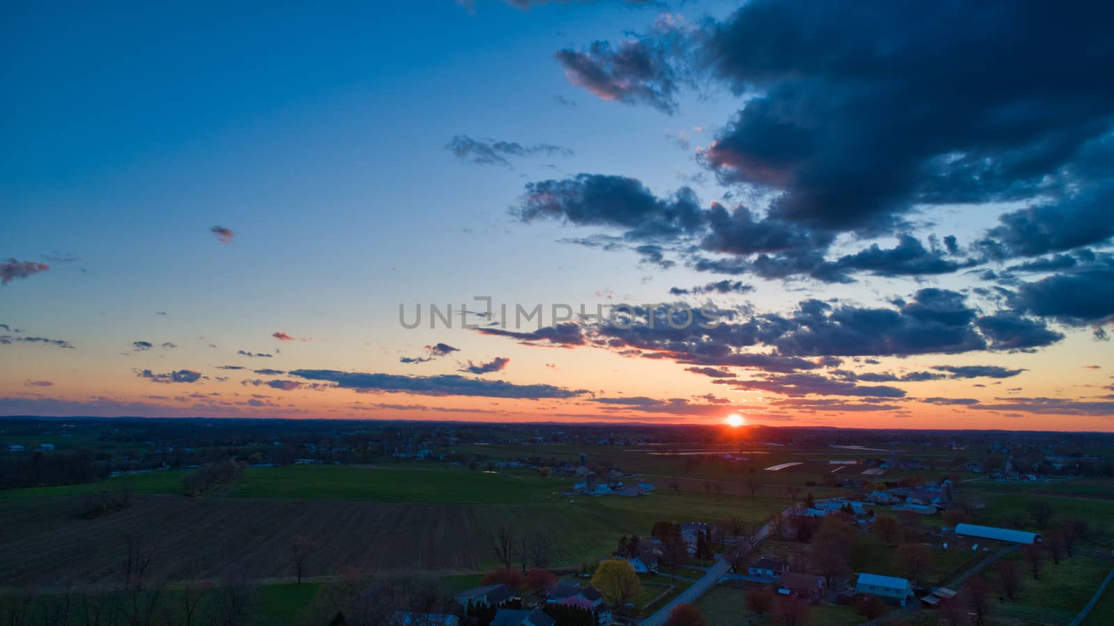 Aerial view of a Sunset over barns, silos and farmlands during the golden hour with blue sky, clouds and red sun