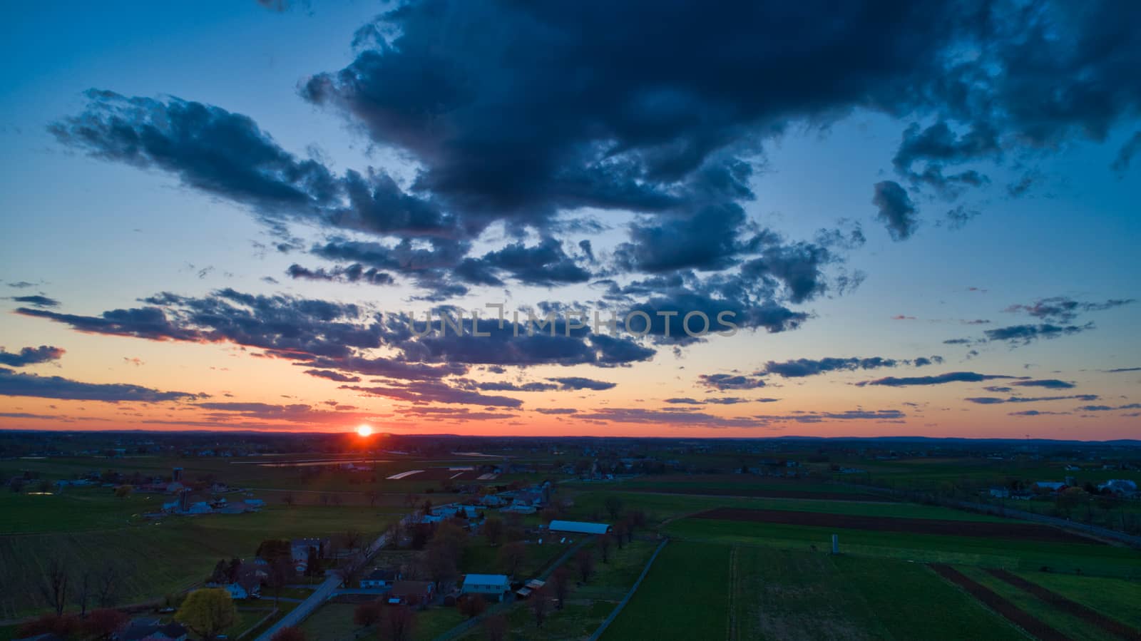 Aerial view of a Sunset over barns, silos and farmlands during the golden hour with blue sky, clouds and red sun