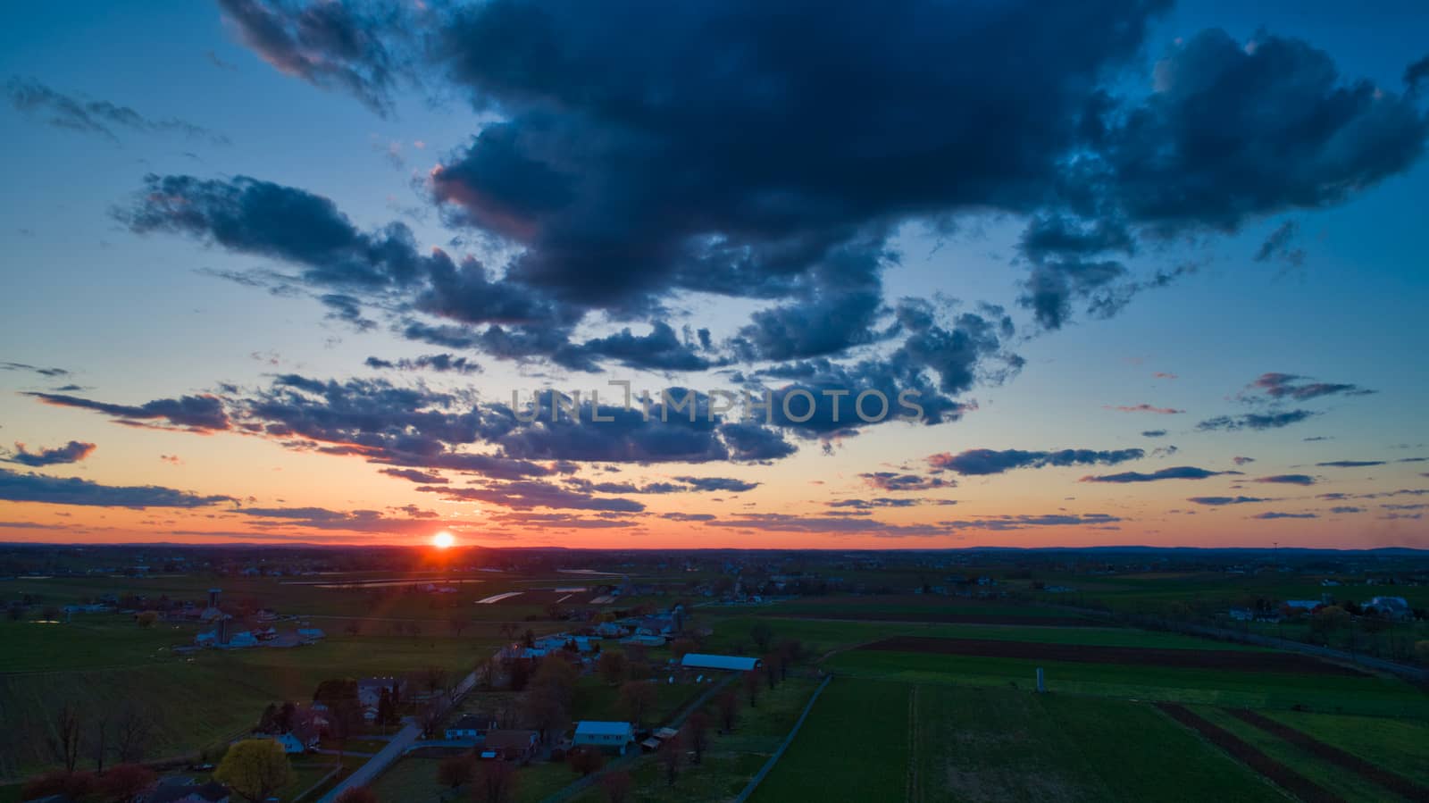 Aerial view of a Sunset over barns, silos and farmlands during the golden hour with blue sky, clouds and red sun