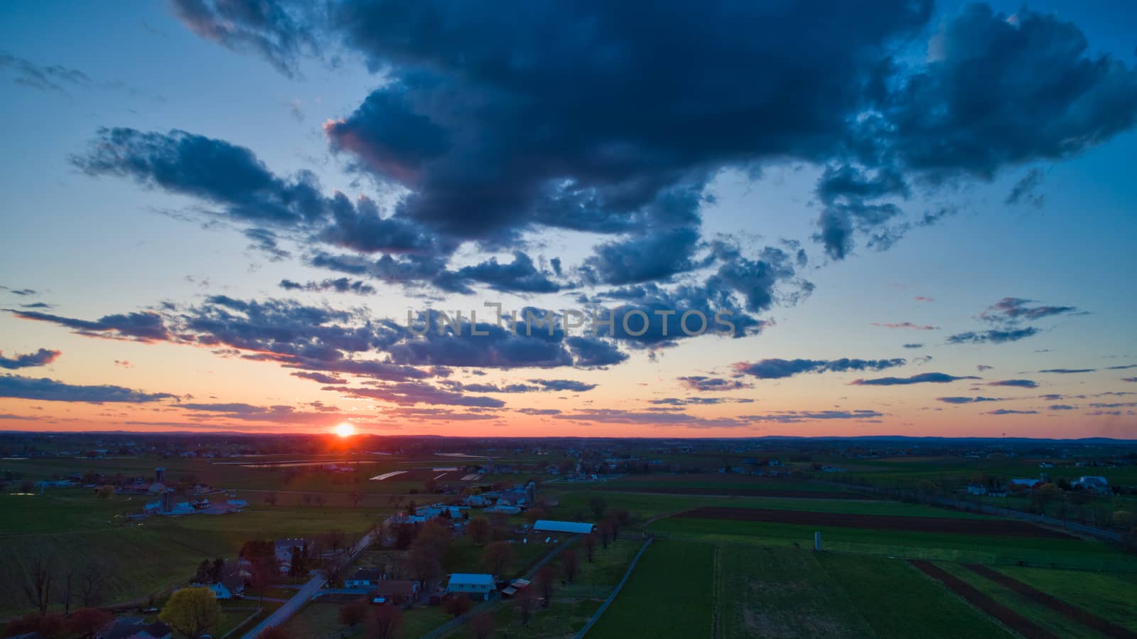 Aerial view of a Sunset over barns, silos and farmlands during the golden hour with blue sky, clouds and red sun