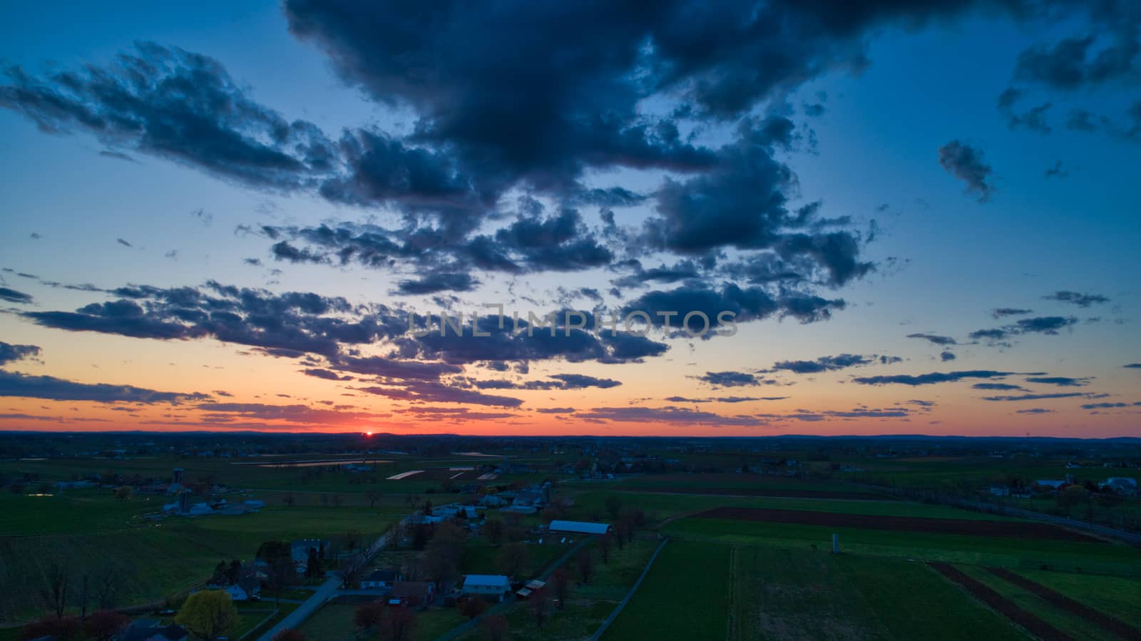 Aerial view of a Sunset over barns, silos and farmlands during the golden hour with blue sky, clouds and red sun