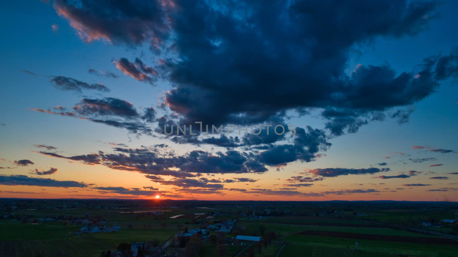 Aerial view of a Sunset over barns, silos and farmlands during the golden hour with blue sky, clouds and red sun