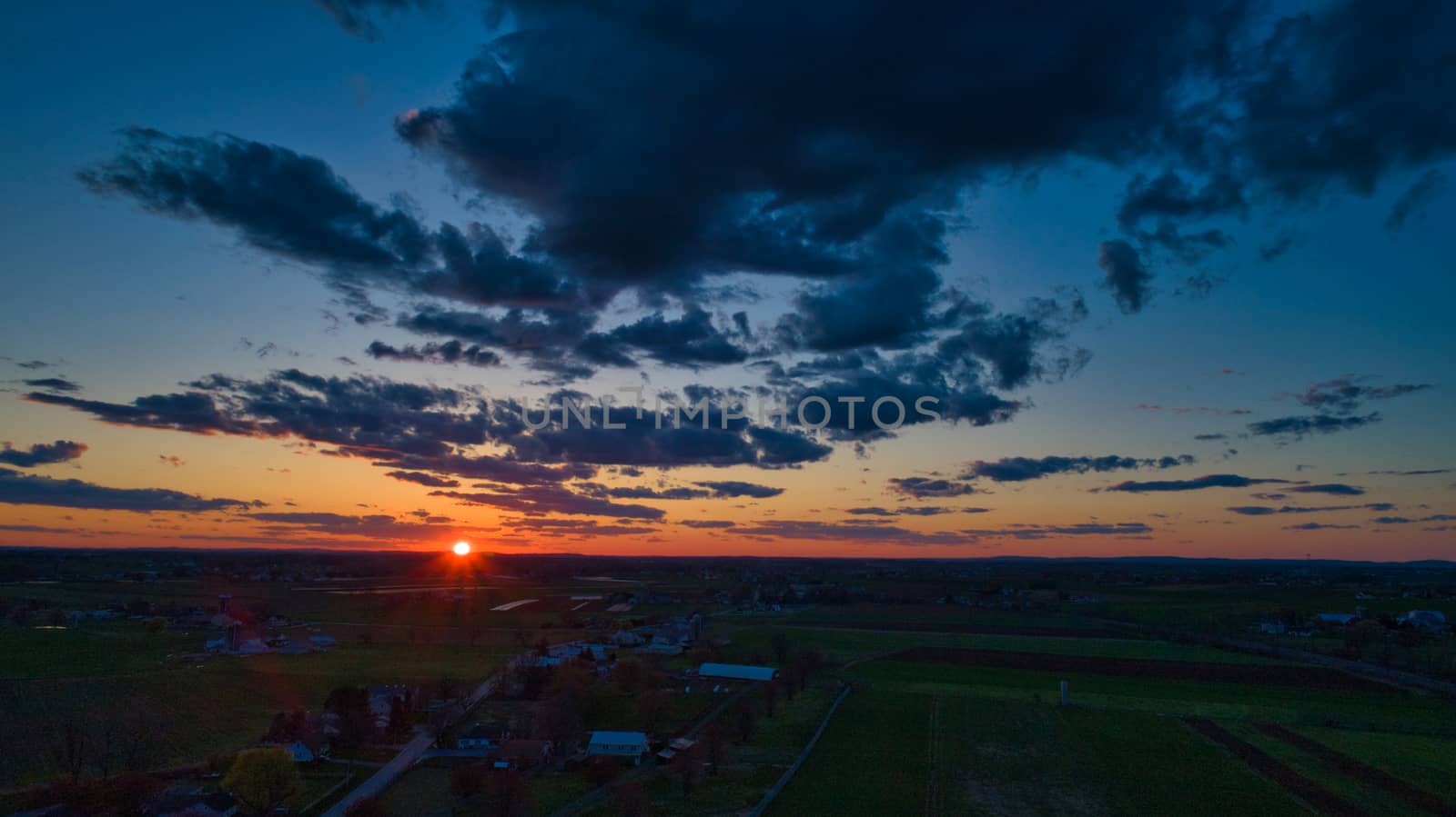 Aerial view of a Sunset over barns, silos and farmlands during the golden hour with blue sky, clouds and red sun