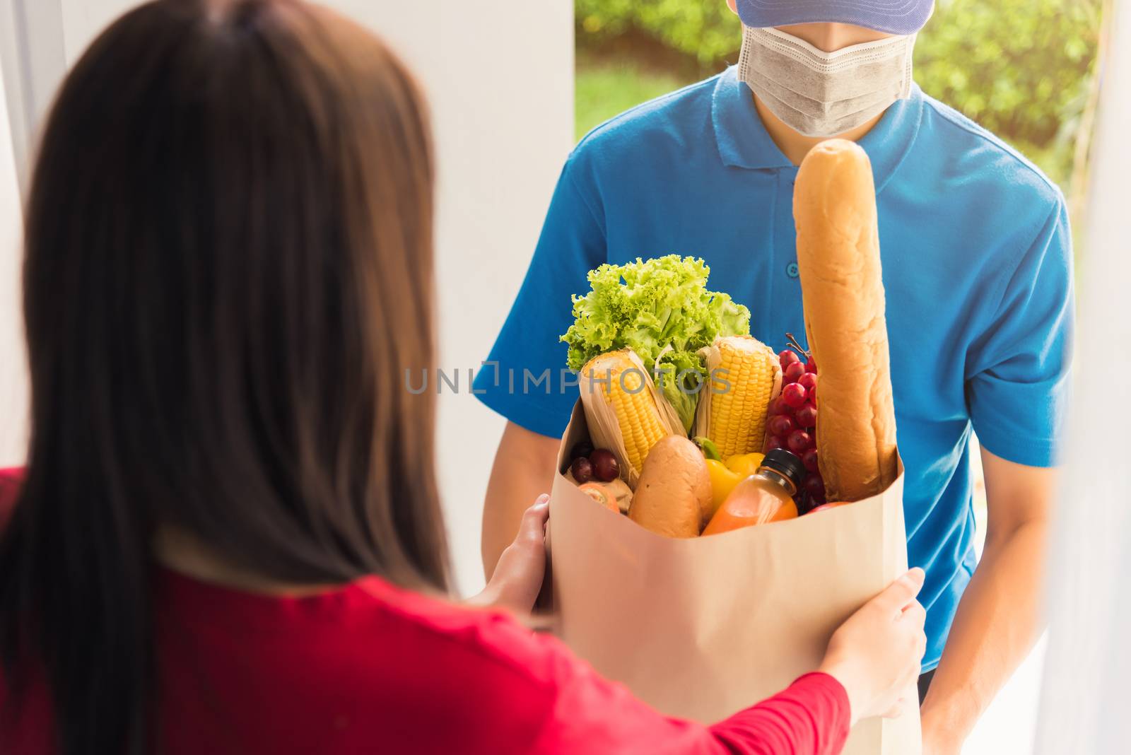 Asian young delivery man in uniform wear protective face mask he making grocery service giving fresh food to woman customer receiving front house under pandemic coronavirus, Back to new normal concept