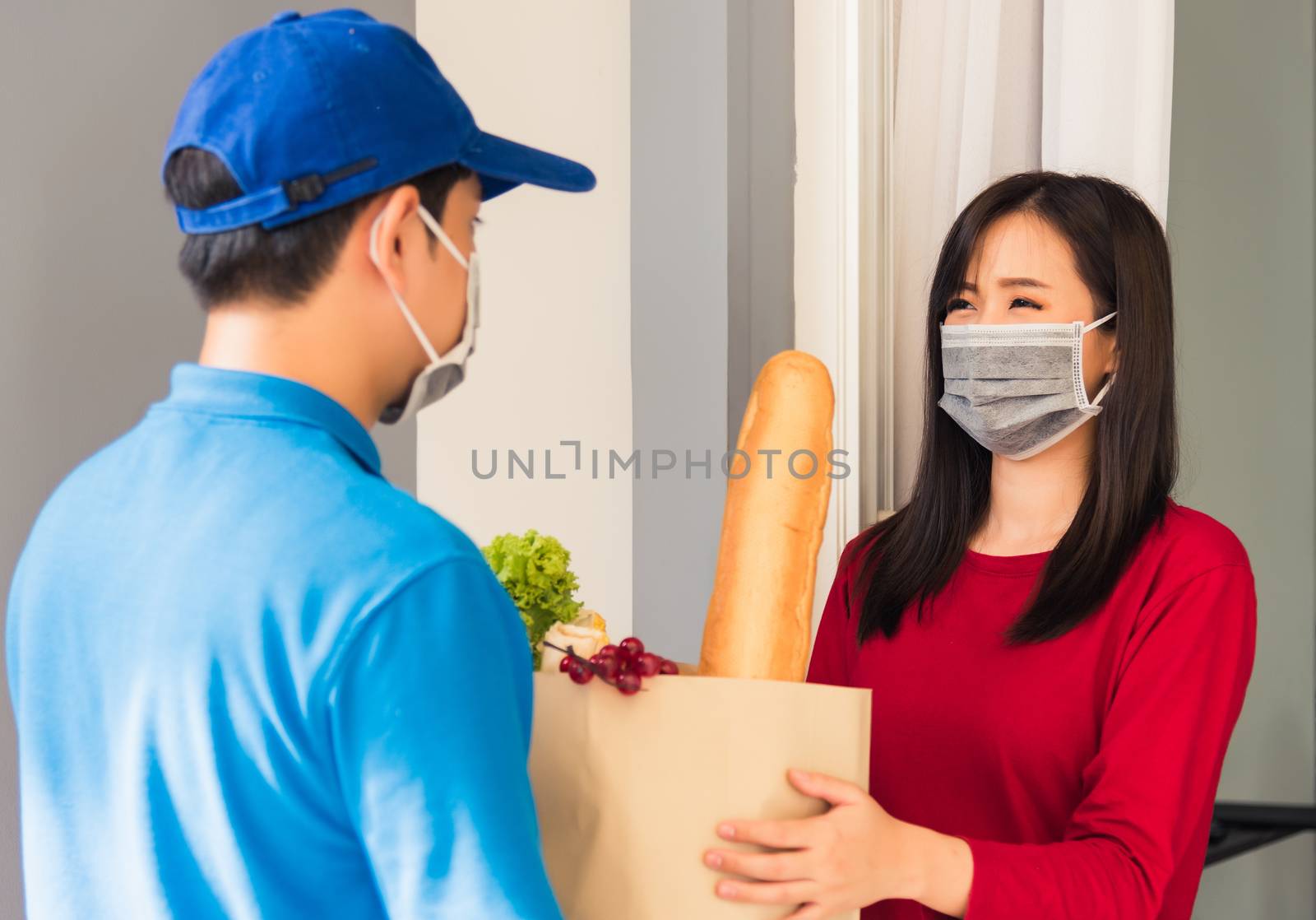 Asian young delivery man in uniform wear protective face mask he making grocery service giving fresh food to woman customer receiving front house under pandemic coronavirus, Back to new normal concept