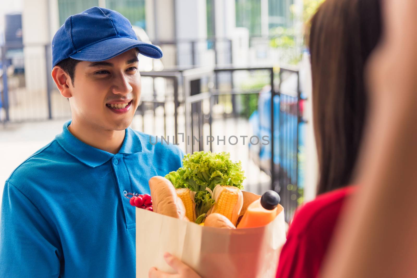 Asian young delivery man in uniform making grocery fast service giving fresh food in paper bag to woman customer receiving at house door under pandemic coronavirus, Back to new normal concept