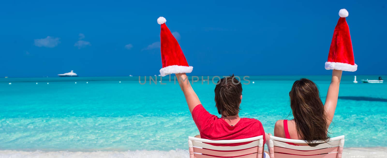 Happy young couple in red Santa Hats sitting on beach chairs by travnikovstudio