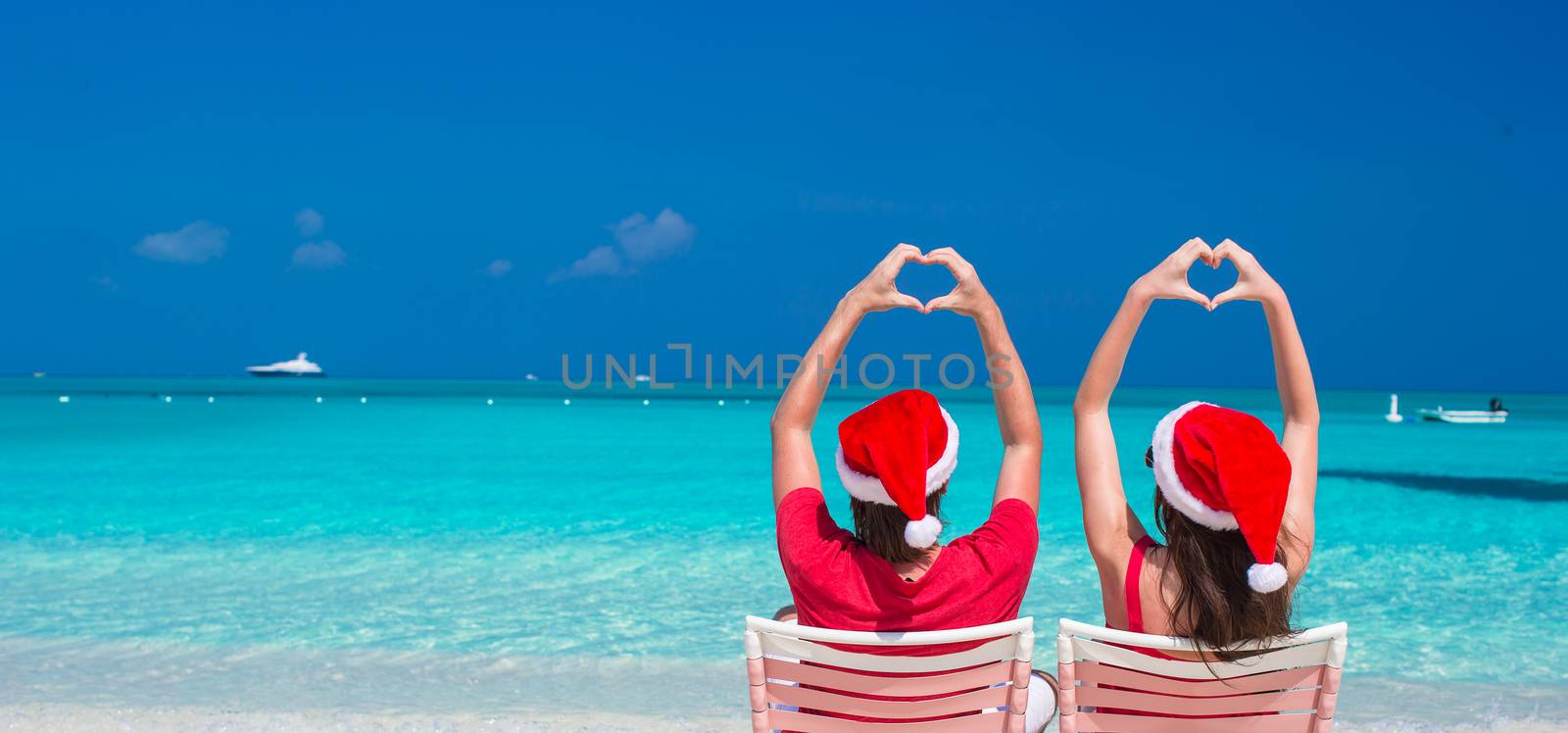 Happy romantic couple in red Santa Hats at beach making hearts by travnikovstudio