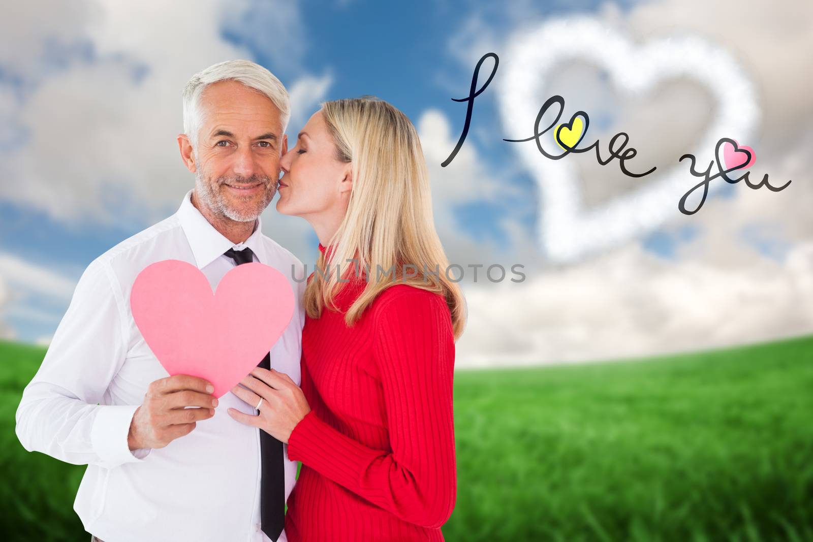 Handsome man holding paper heart getting a kiss from wife against green field under blue sky