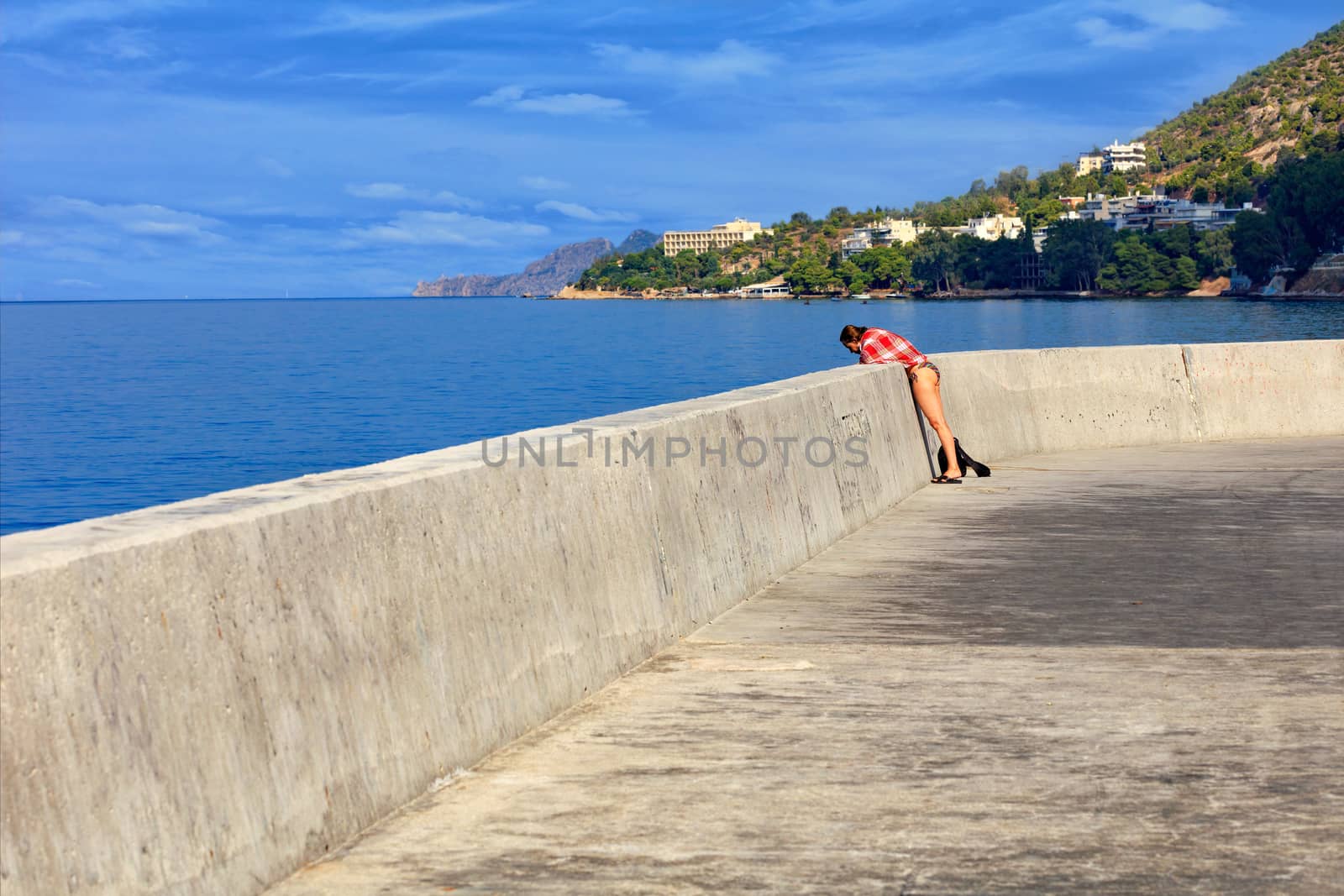 On the concrete pier of the sea bay, a lady in a beach suit observes the life of marine coastal fish and inhabitants against the backdrop of a receding perspective of the sea coastline and the hillside, image with copy space.