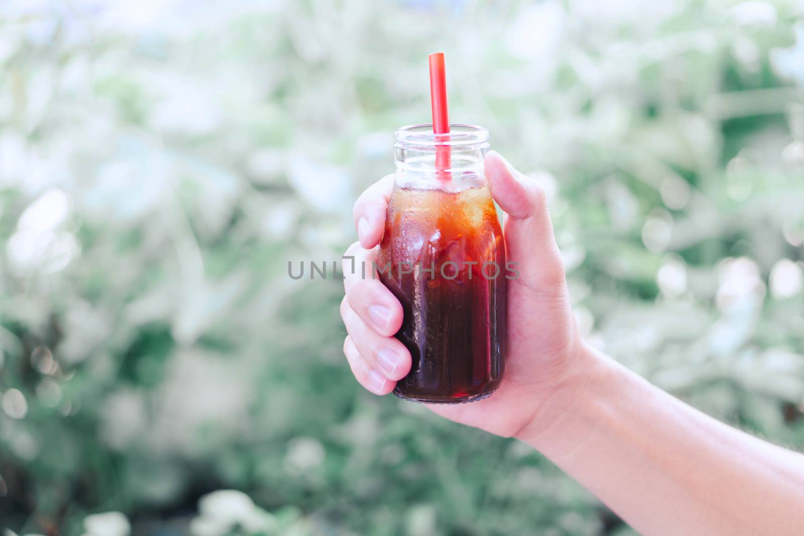 Close up hand holding bottle glass of americano coffee with green nature background, selective focus