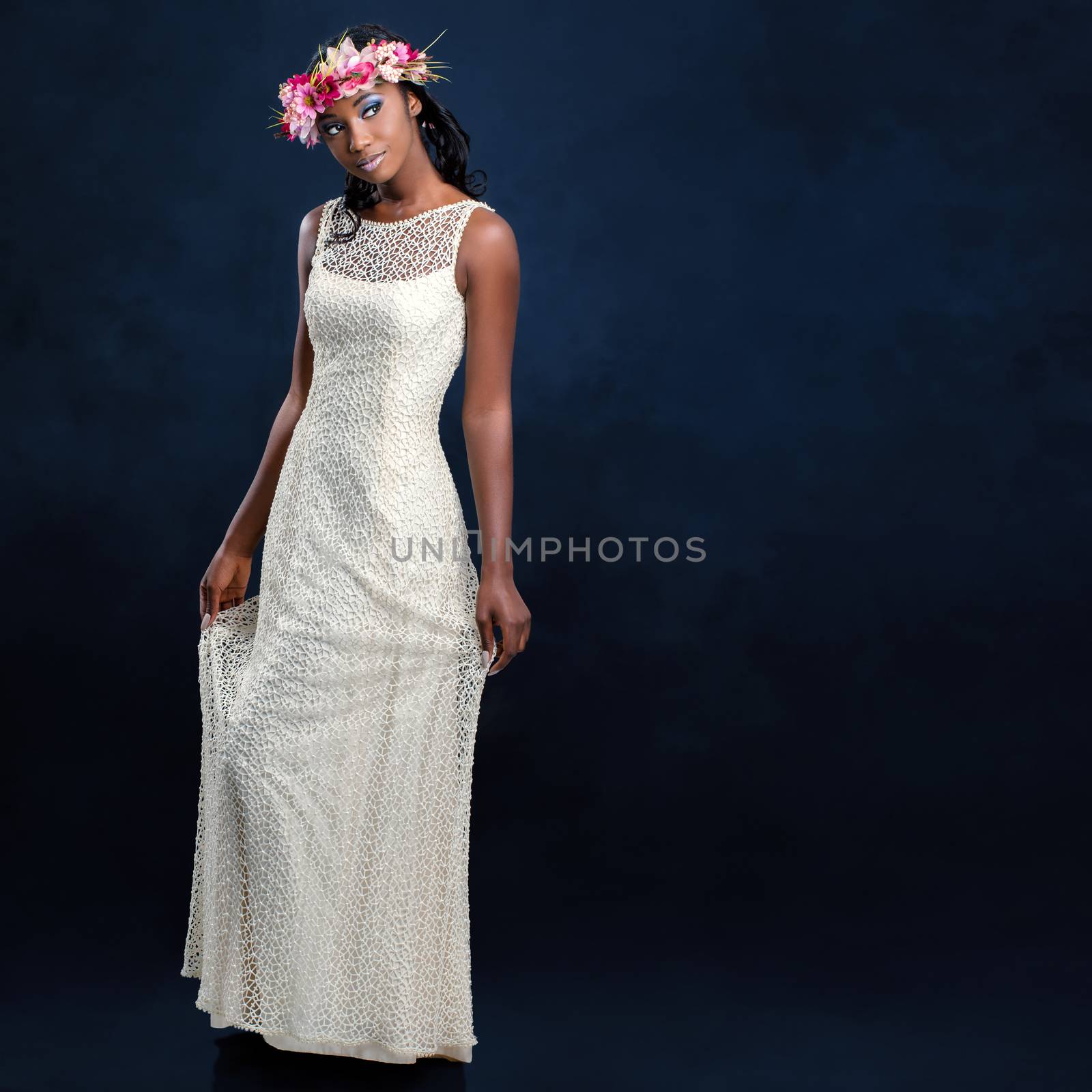 Full length studio portrait of beautiful young african bride in long white wedding gown. Woman wearing colorful flower garland against dark blue background.