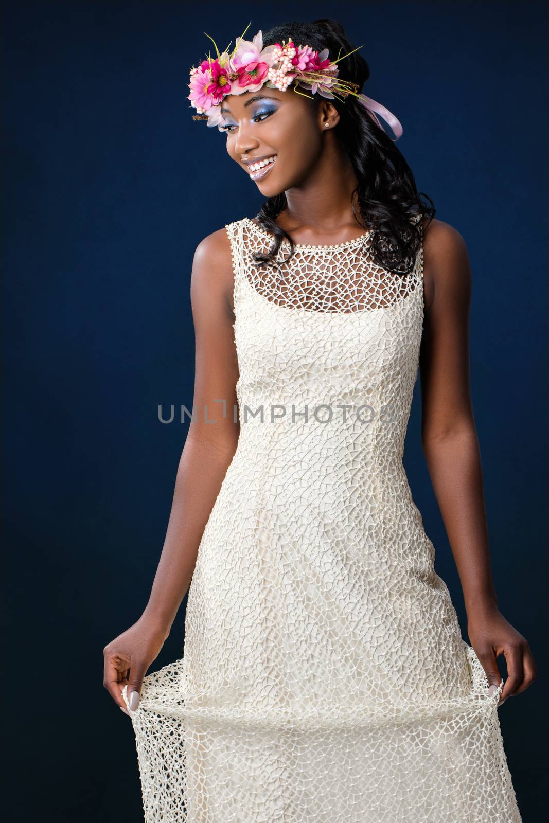 Close up studio portrait of attractive young african bride wearing wedding gown and colorful flower wreath.Laughing girl looking aside against dark blue background.