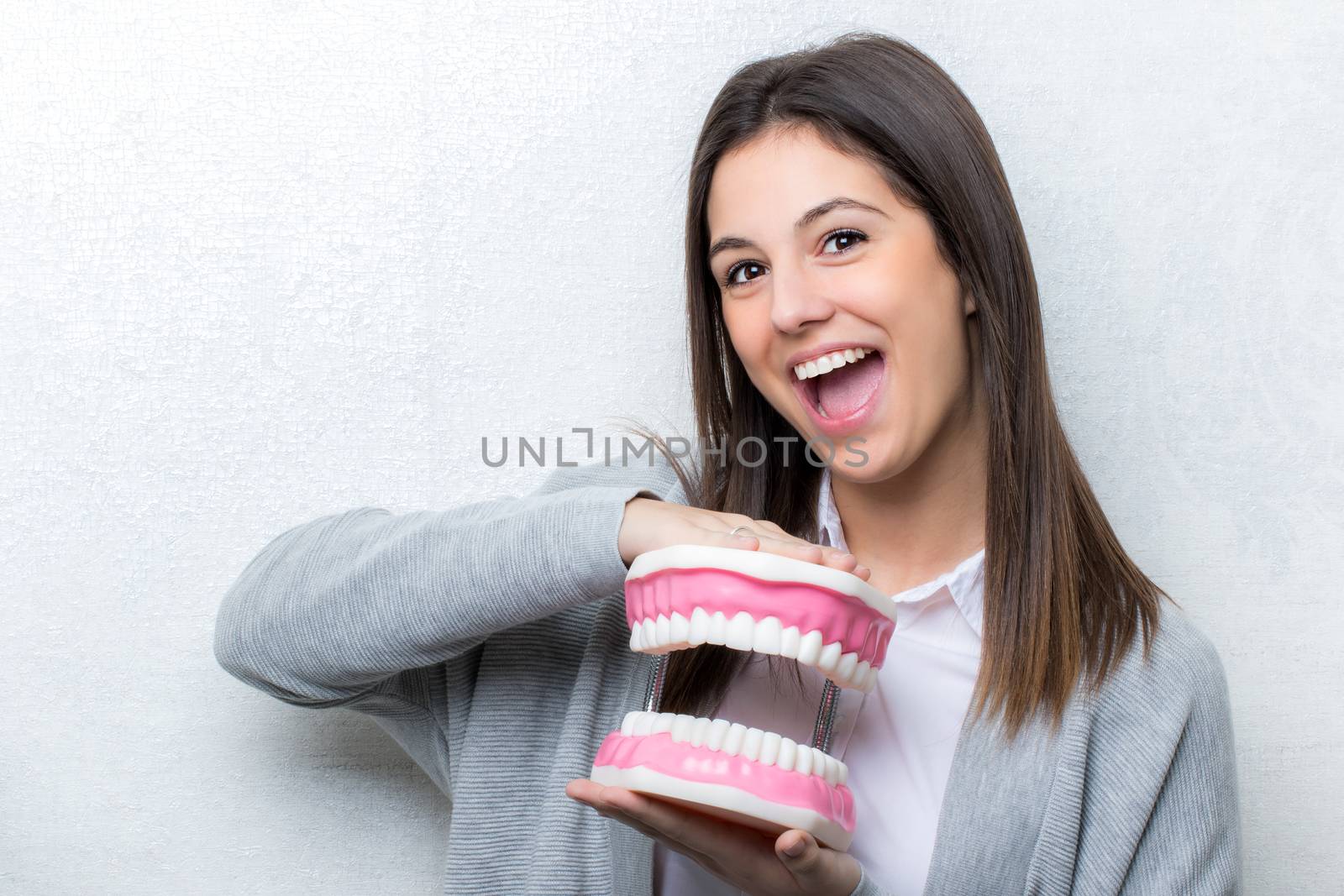 Close up portrait of attractive young girl holding oversize human teeth prosthesis.Woman  with open mouth facial expression against seamless light background.