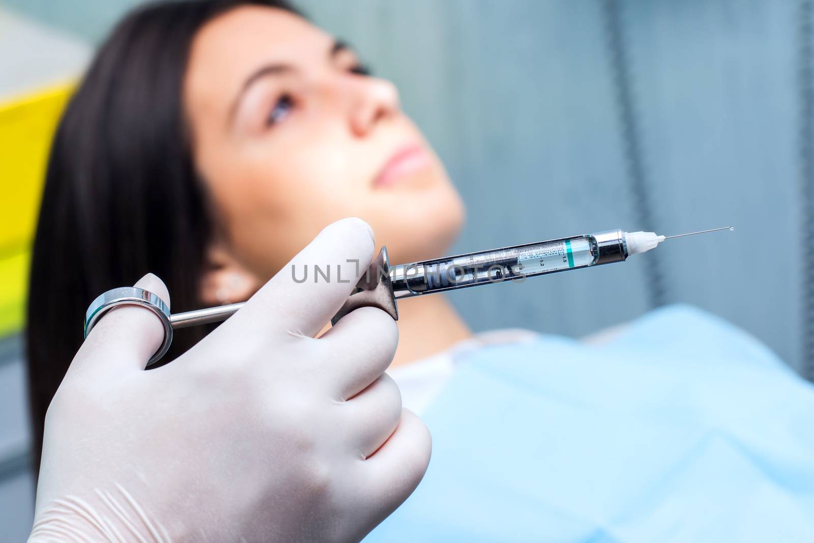 Macro close up of hand with glove holding medical syringe with out of focus patient in background.