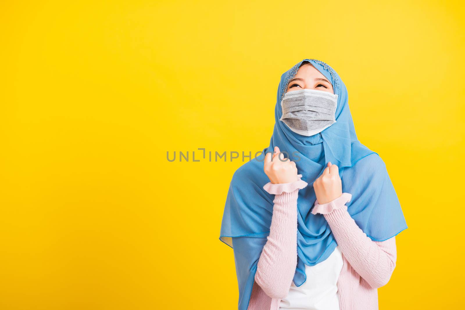 Muslim Arab, Happy beautiful young woman wear veil hijab wear protective face mask quarantines coronavirus raises hands glad excited cheerful after recovering from illness, isolated yellow background