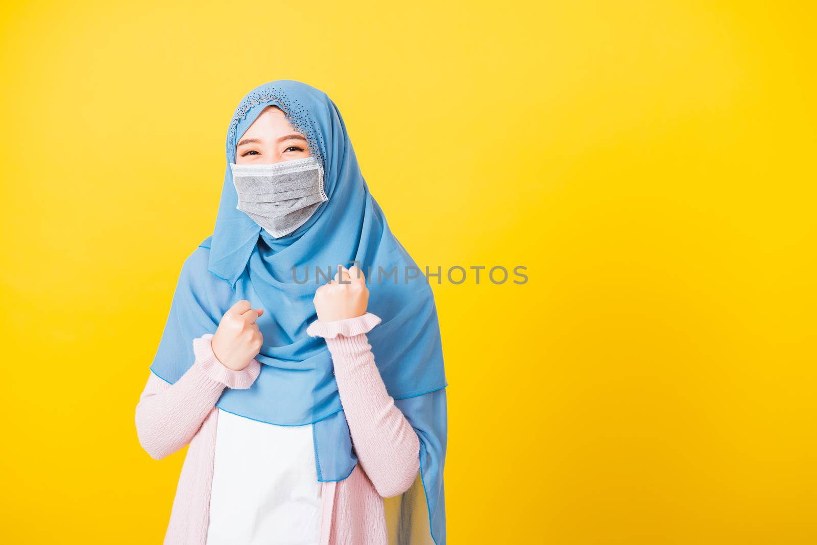 Muslim Arab, Happy beautiful young woman wear veil hijab wear protective face mask quarantines coronavirus raises hands glad excited cheerful after recovering from illness, isolated yellow background