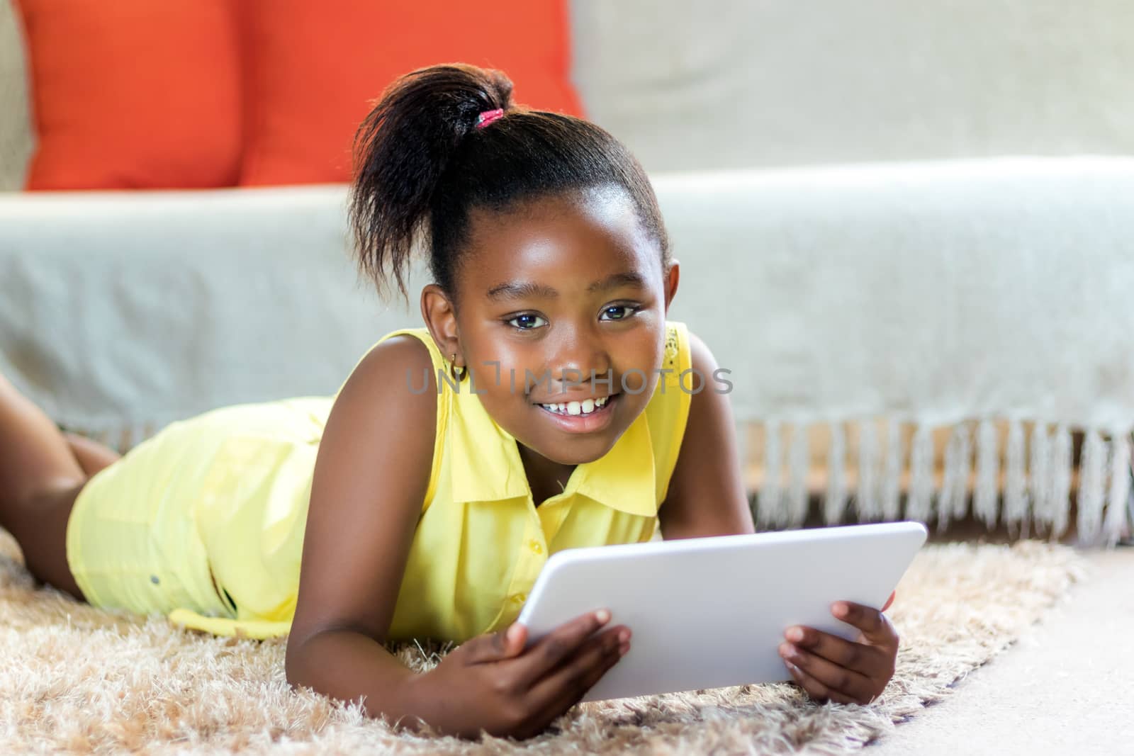 Close up portrait of cute little african girl with ponytail laying on carpet in lounge with tablet.
