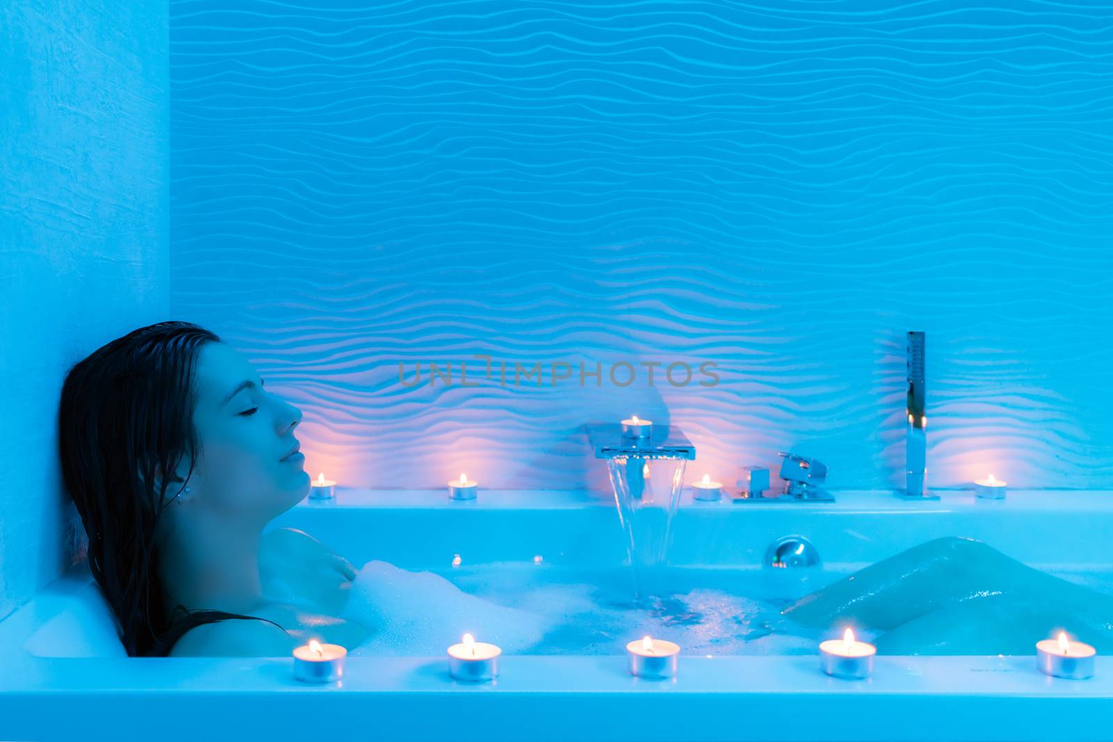 Close up portrait of young woman relaxing in hot foam bath at home.Low key blue ambient with decorative candles along bath tub.