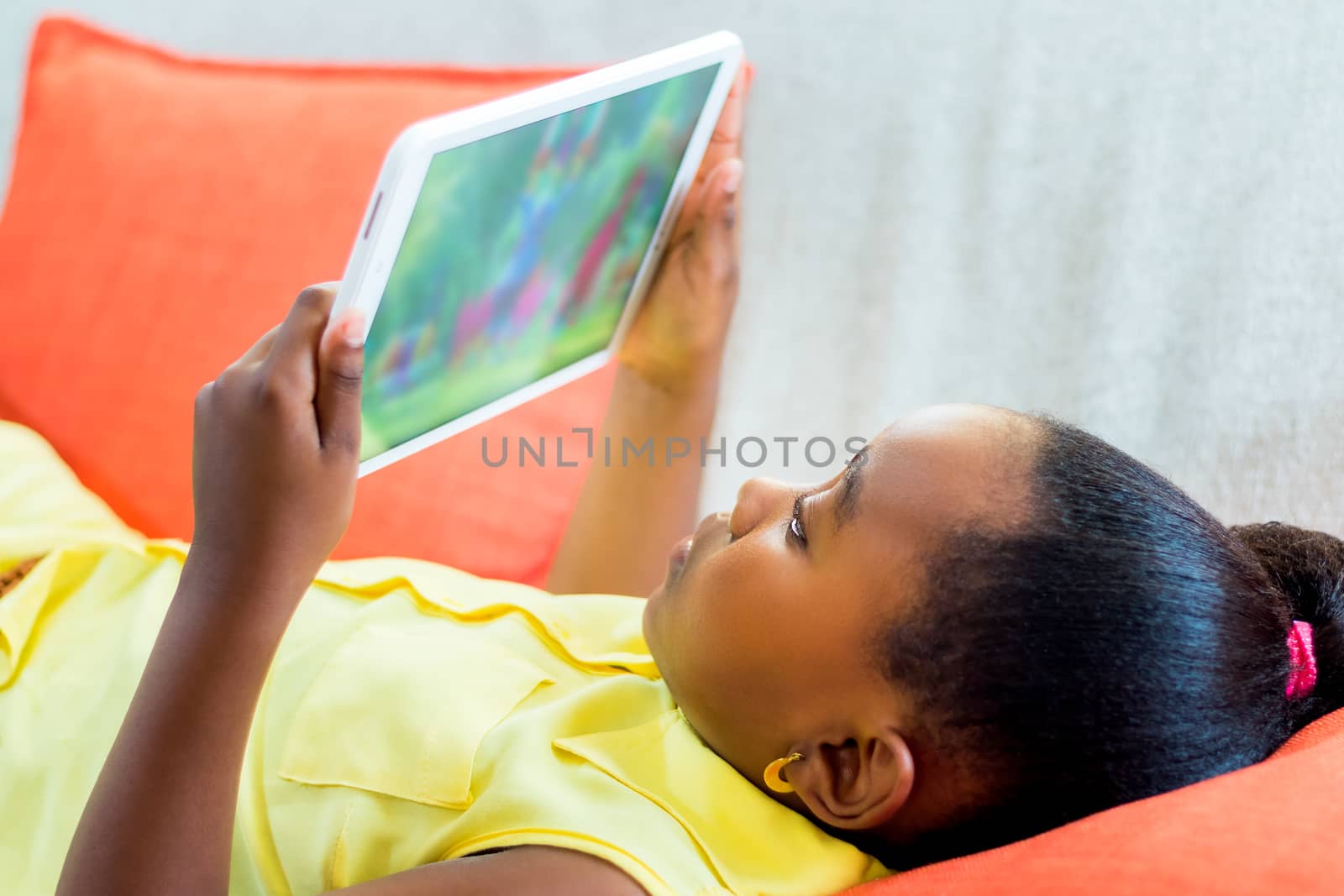 Close up portrait of cute little african girl playing with digital tablet. Kid laying on couch looking at movie.