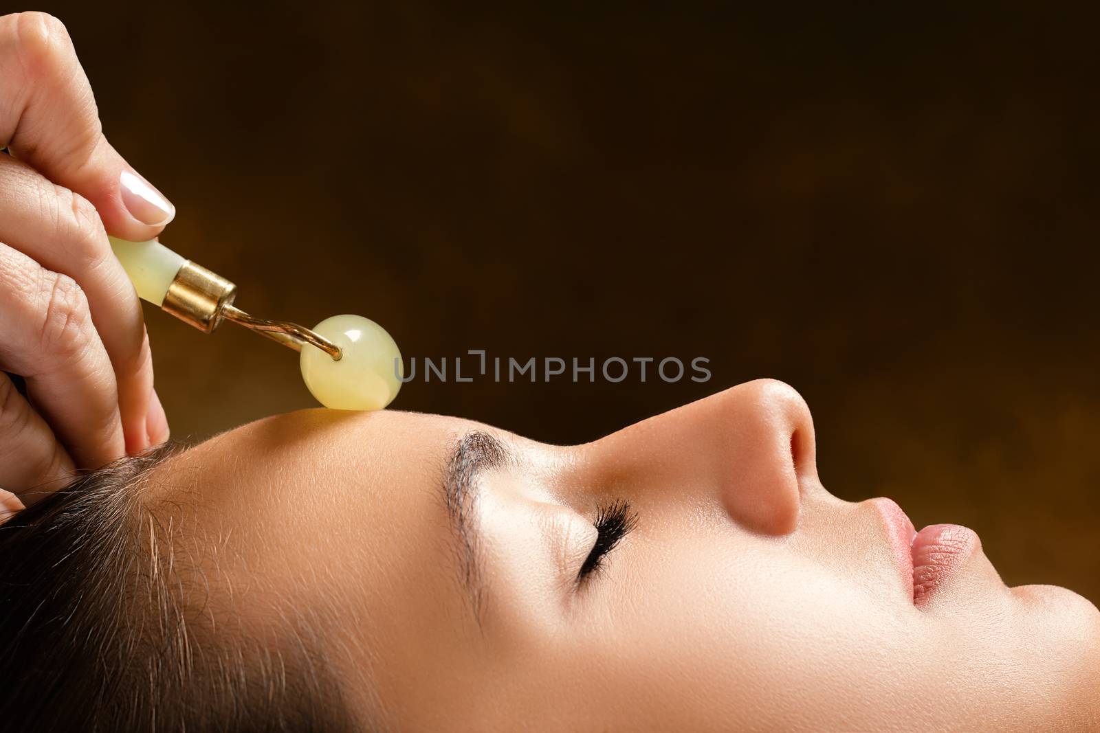 Macro close up side view of woman having beauty treatment in spa.Therapist applying jade roller on forehead.