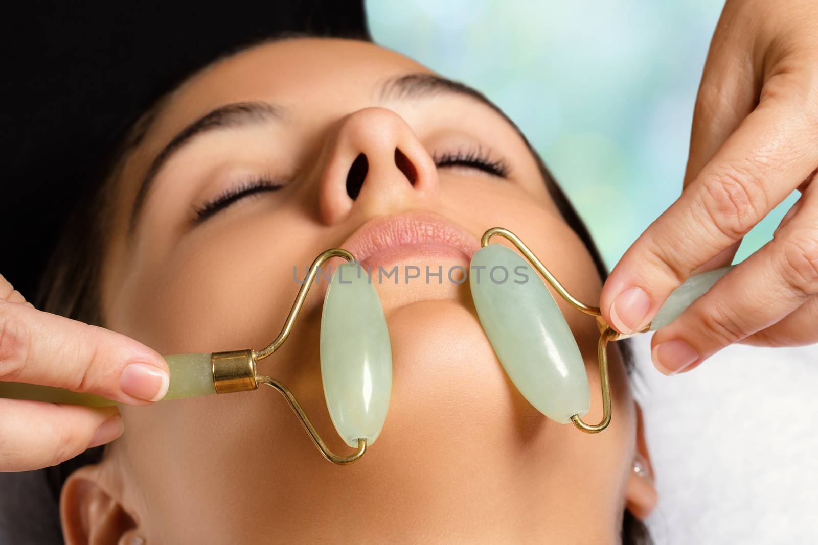 Macro close up portrait of woman having facial beauty treatment in spa. Therapist massaging chin with jade rollers.
