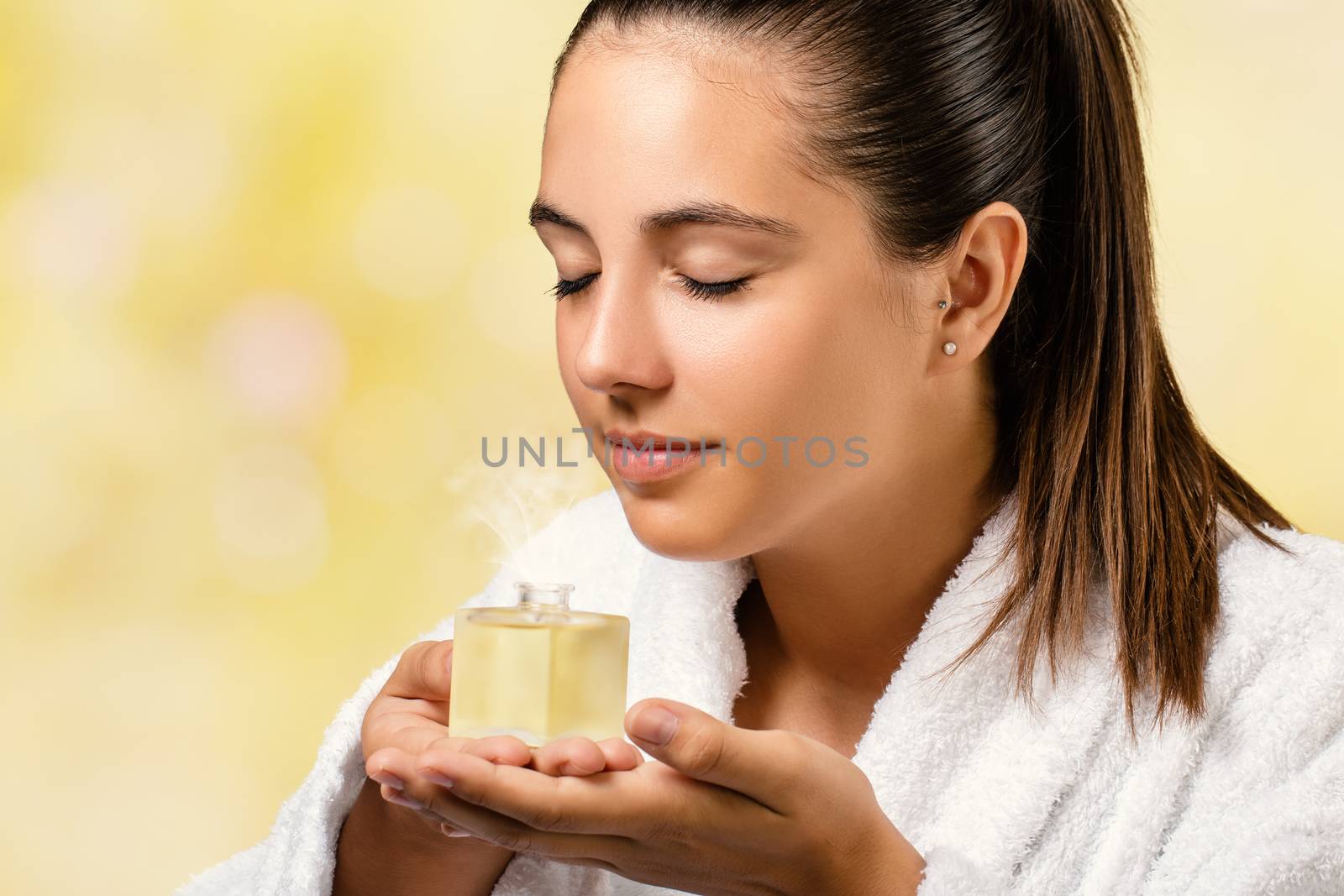 Close up portrait of woman in white spa gown smelling essential oil fragrance.Girl holding little bottle with aromatic scent.