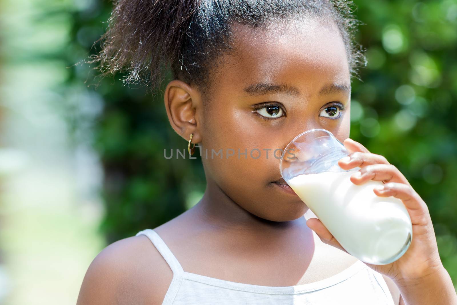 Close up portrait of cute little african girl drinking glass of milk in garden.