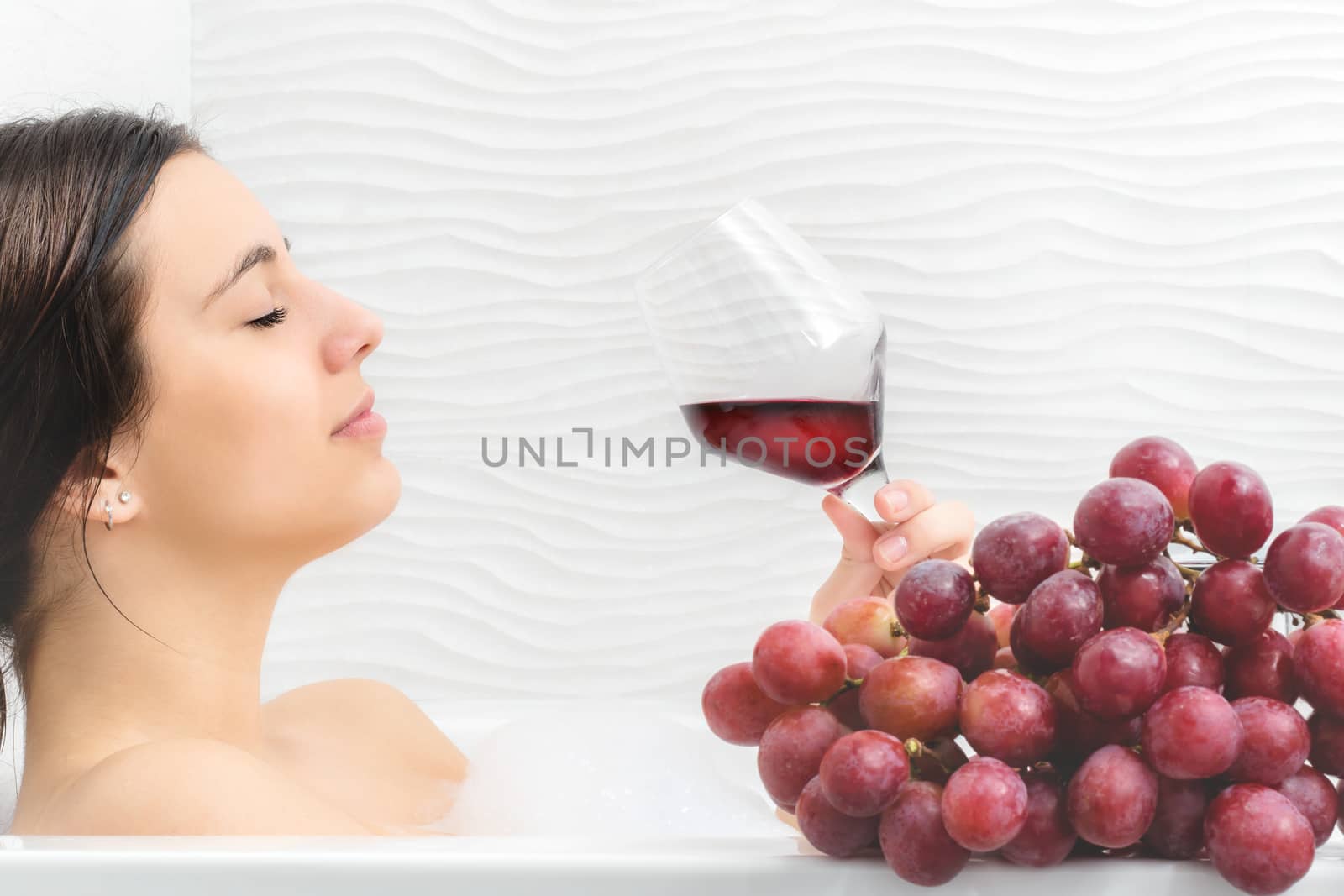 Close up portrait of young woman relaxing in foam bath with red wine and bunch of red grapes.Girl holding glass with red wine. 