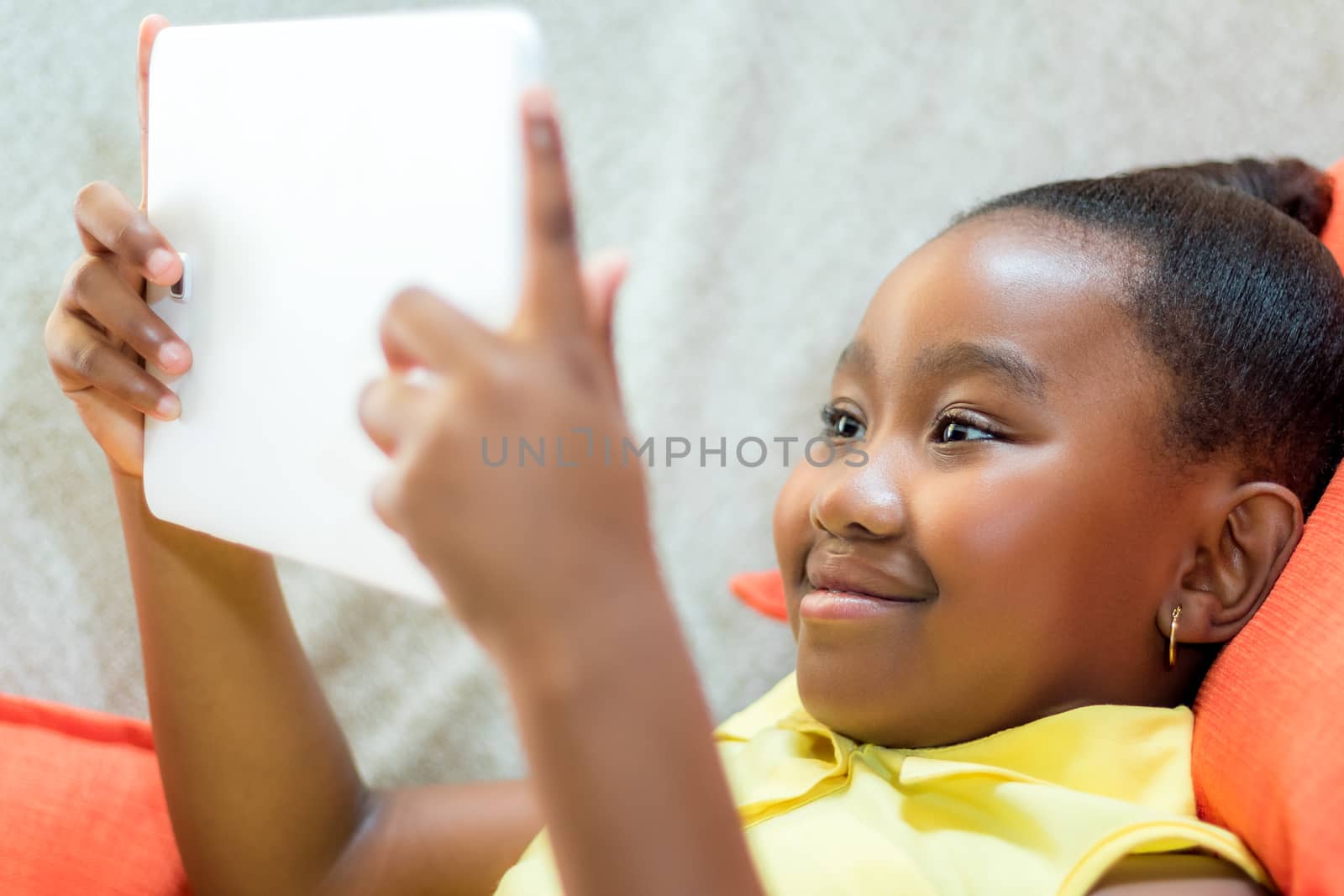 Close up portrait of cute little african girl playing with digital tablet. Kid laying on couch looking at movie.